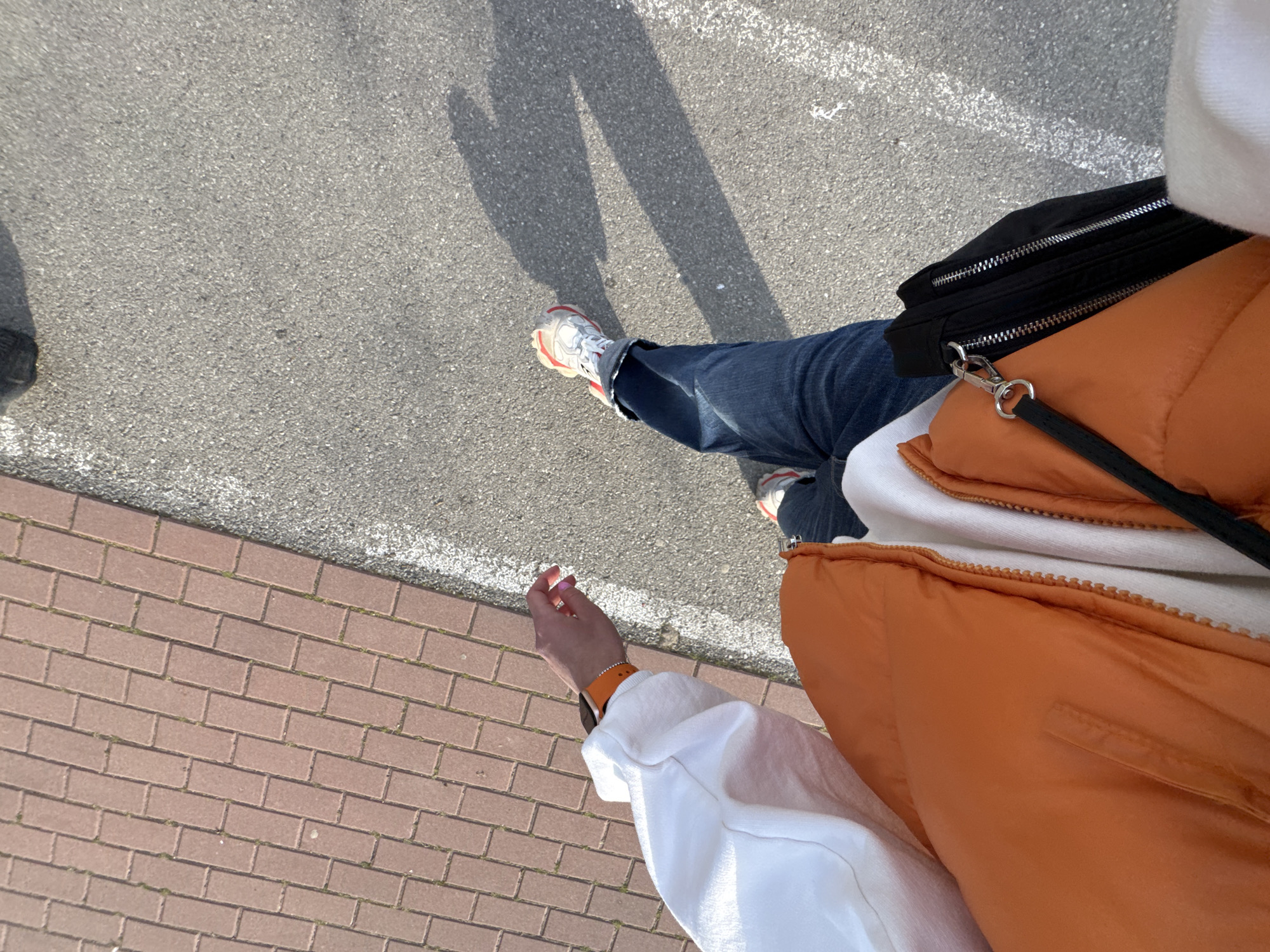 The image shows a first-person perspective of someone standing on a paved surface next to a brick sidewalk. The paved surface has a white line. The person is wearing blue jeans, white and red sneakers, an orange puffer vest over a white hooded sweatshirt, and a black fanny pack. The person has an orange smartwatch on their left wrist. The image's composition does not provide sufficient context to determine a specific location in Blagoevgrad, Bulgaria. The dominant subjects include the person's clothing, accessories, and the surrounding pavement and sidewalk.
