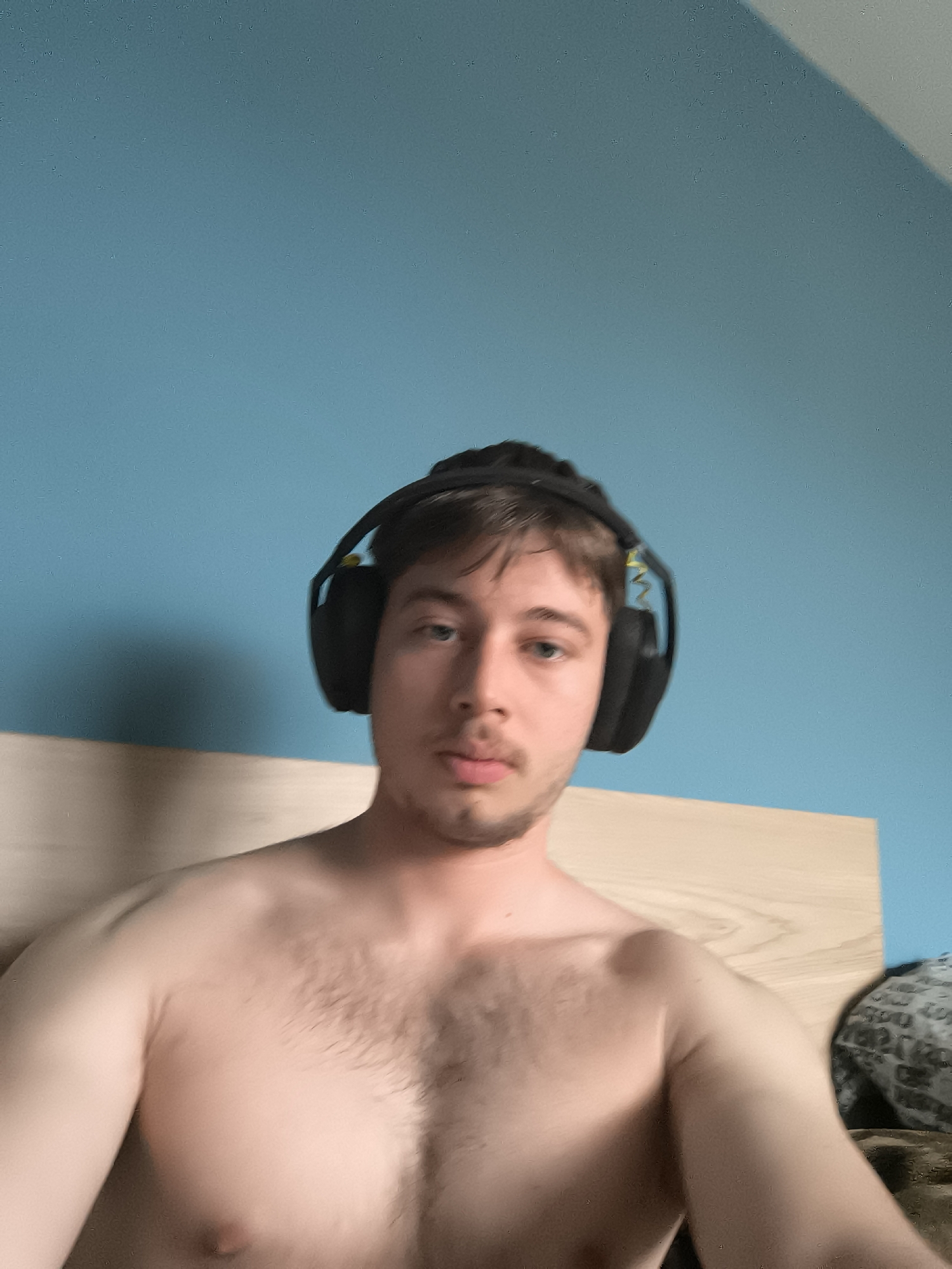 A shirtless man wearing black headphones is centered in the frame. He has fair skin, brown hair, and a light beard. Behind him is a light-colored wooden headboard and a blue wall. Part of a patterned pillow is visible in the lower right corner. The location is indeterminable from the image and no evidence suggests it is Sofia, Bulgaria.
