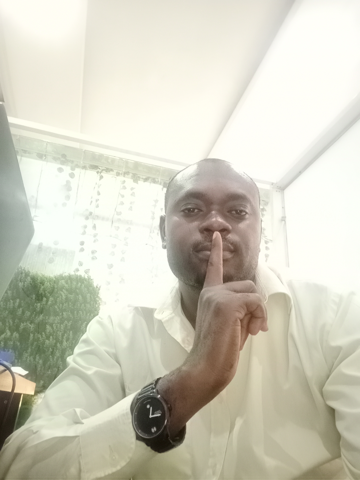A medium shot depicts a dark-skinned man holding his index finger to his lips in a "shush" gesture. He is wearing a light-colored button-down shirt and a black wristwatch on his left wrist. Behind him, a window provides a view of greenery, possibly foliage or bushes. The lighting appears artificial and bright. The setting is likely indoors, given the presence of ceiling fixtures. No specific details directly indicate the location in Gwarinpa, Nigeria; the setting could be an office or residence but its location in Nigeria is unknown.