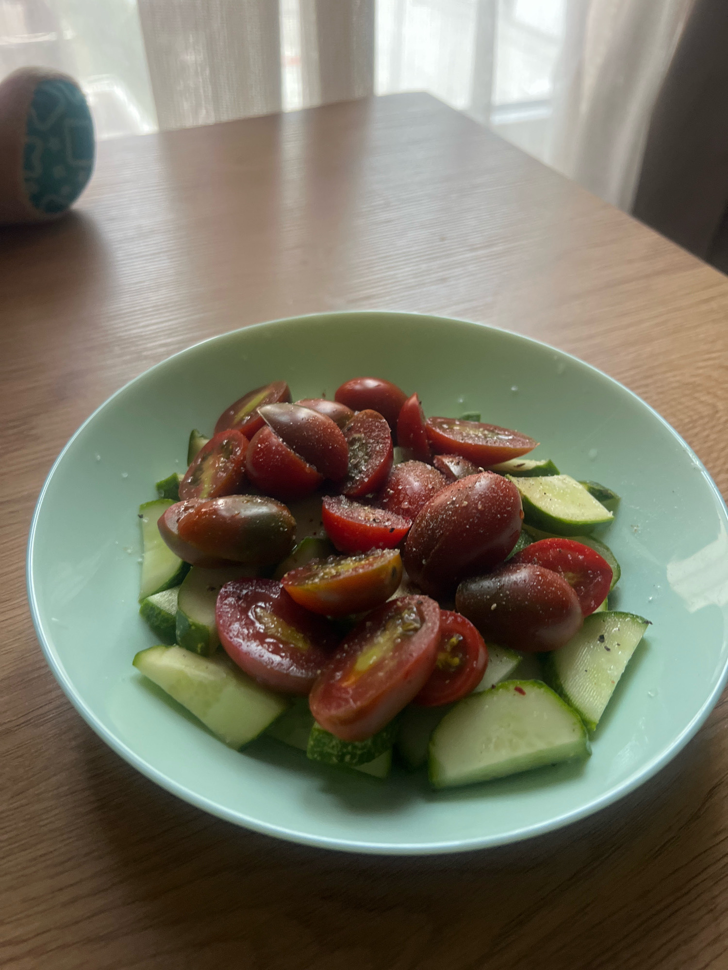 A close-up, eye-level shot captures a light green bowl filled with a salad on a wooden table. The salad consists of halved cherry tomatoes, some appearing dark red and others with lighter red and yellow hues, mixed with sliced cucumbers. The vegetables are seasoned with salt and pepper. The wooden table has a visible grain pattern. In the upper left background, a blurred object with a blue and green patterned end is visible, and a sheer white curtain is present in the far background. The scene is indoors, with soft lighting suggesting natural light coming from a window. The image does not provide any information about the city of Plovdiv, Bulgaria, nor does it contain any people or indicate any actions or interactions.