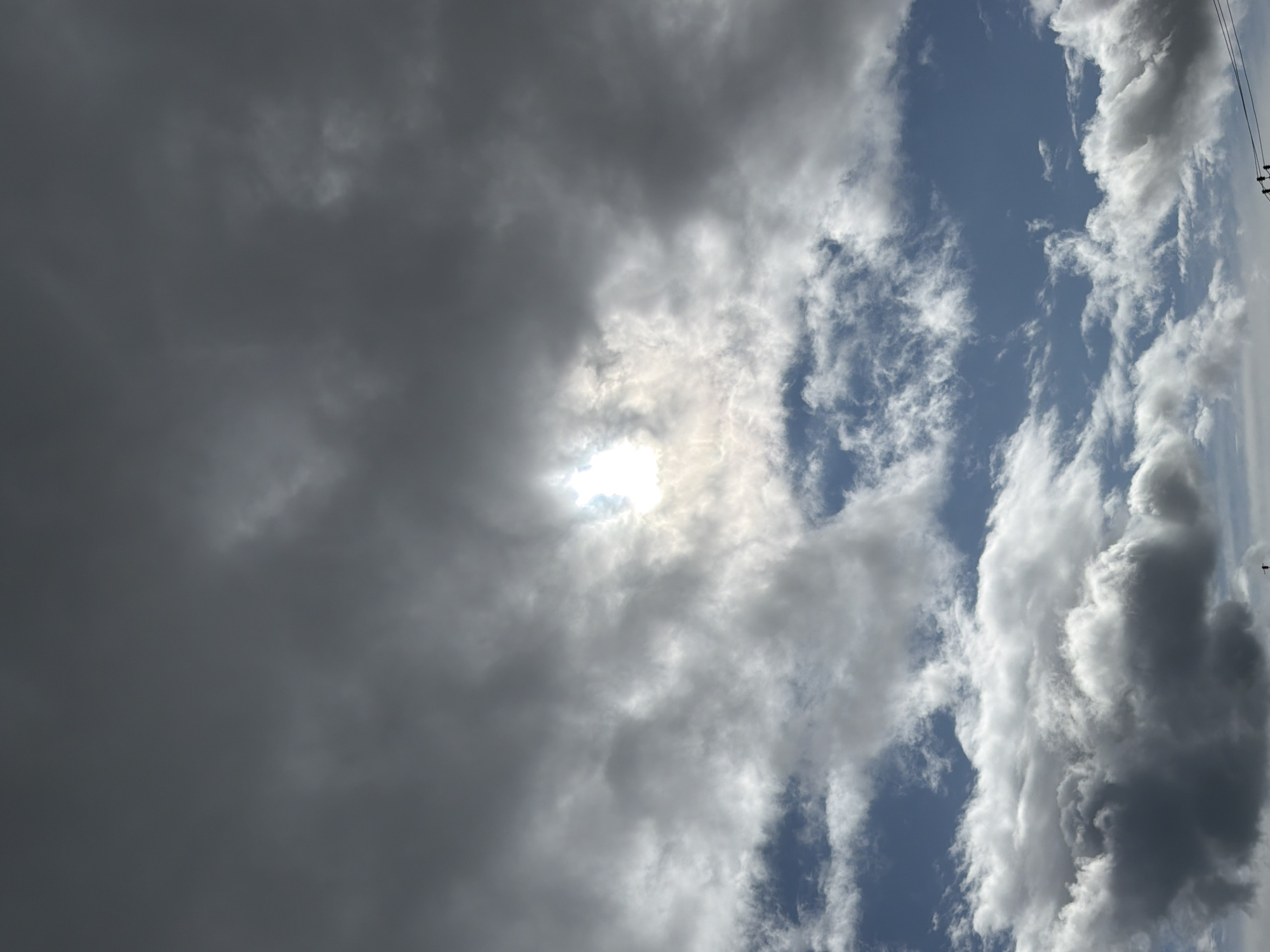 The image shows a sky with dramatic cloud formations. The sun is partially obscured by clouds, creating a bright, hazy glow in the center of the frame. Darker, heavier clouds dominate the upper portion of the image, while lighter, more wispy clouds are visible lower down against a blue sky. The overall impression is of a dynamic and overcast sky. There are no discernible buildings, people, or specific landmarks visible in this sky-focused image. The image does not contain any information that could place it in Yuzhno-Kurilsk, Russia.