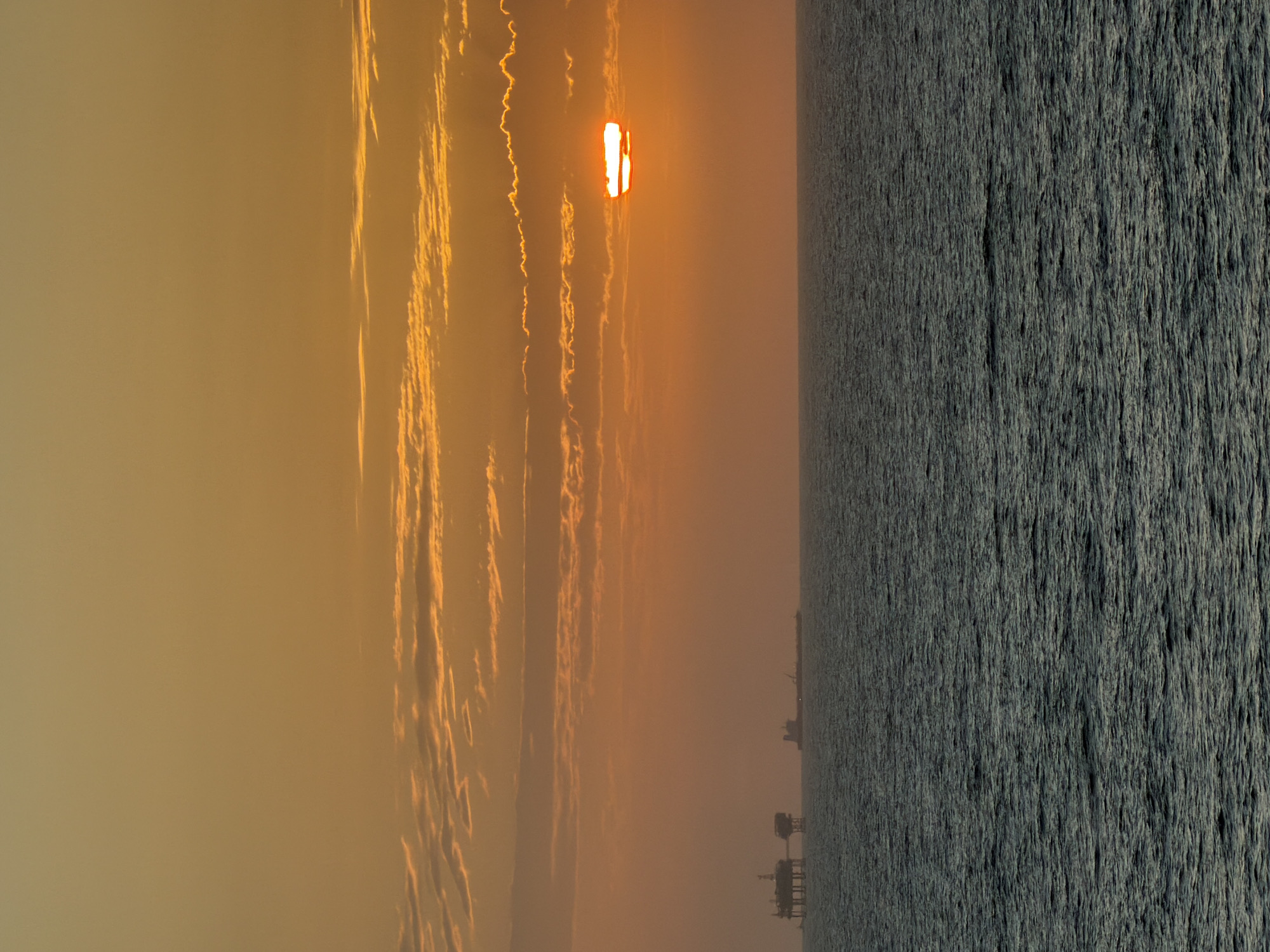 The image captures a sunset or sunrise over a vast body of water, likely the sea or ocean. The sky is dominated by warm, golden-orange and yellow hues, with a bright, elongated sun appearing near the center, partially obscured by thin, wispy clouds that stretch horizontally across the frame. These clouds are illuminated with intense orange and yellow light, creating striking patterns.
The lower half of the image features a dark, textured expanse of water, appearing deep blue-grey, indicating small waves or ripples on its surface. Along the distant horizon line, several indistinct structures, possibly oil rigs or offshore platforms, are silhouetted against the glowing sky, adding an industrial element to the otherwise natural scene.
The time of day is clearly either early morning or late evening, characterized by the low sun and the golden light saturating the atmosphere. The overall mood is serene yet dramatic, with the hazy air diffusing the sunlight. There is no visible text in the image.