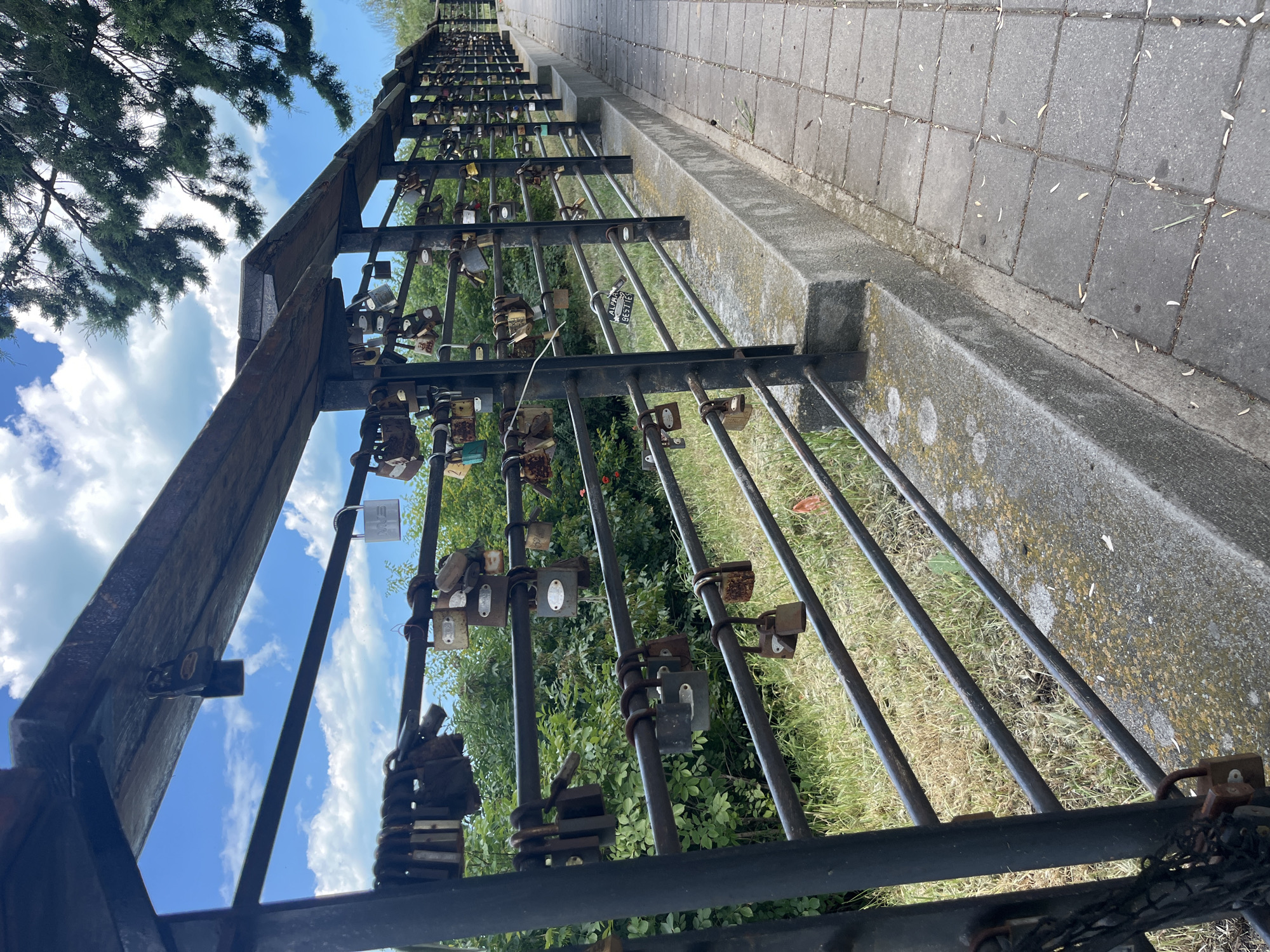 The image depicts a perspective view of a pedestrian walkway alongside a black metal railing on a sunny day. The railing is adorned with numerous padlocks of varying sizes and conditions, many of which appear old and rusted. Behind the railing, lush green foliage and trees are visible, suggesting an outdoor setting. The walkway is paved with grey square tiles. The railing extends into the distance, following the curve of the walkway. The sky is a bright blue with scattered white clouds. This scene is characteristic of the "Bridge of Wishes" or "Love Lock Bridge" located in the Sea Garden of Varna, Bulgaria, a popular spot where couples attach padlocks as symbols of their commitment.