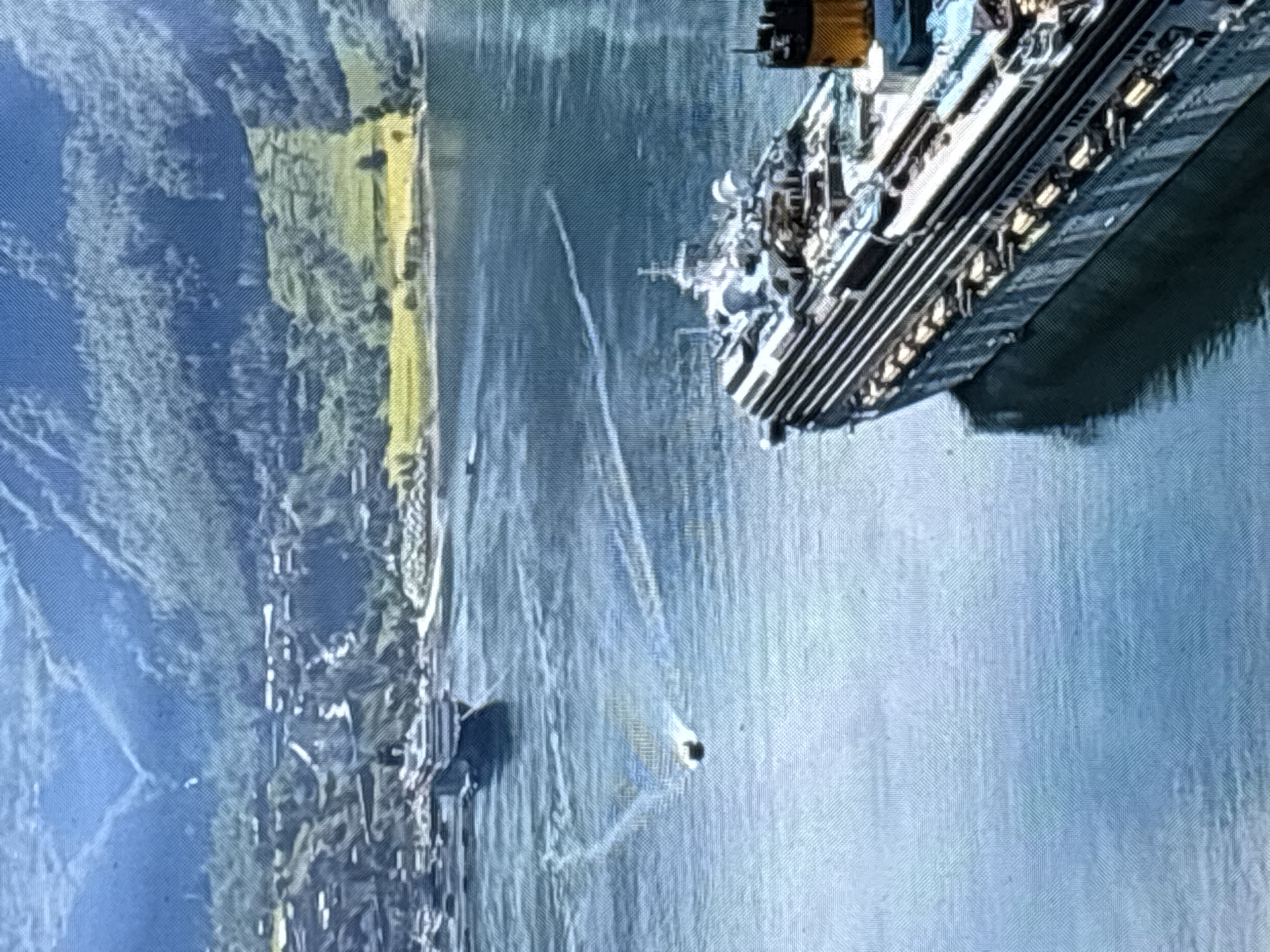 The image displays a panoramic view of a bay with a large cruise ship in the foreground, partially obscuring a smaller vessel in the middle ground. The cruise ship, characterized by its multi-deck structure and white and dark striped hull, is docked or moving slowly on the calm, blue water. In the distance, a mountainous landscape with lush green vegetation and a town nestled along the shore is visible. The town features a cluster of buildings with white roofs, some of which are situated on the slopes of the hills. The overall scene suggests a scenic coastal or fjord-like environment. The image does not depict the city of Zvenigorod, Russia.