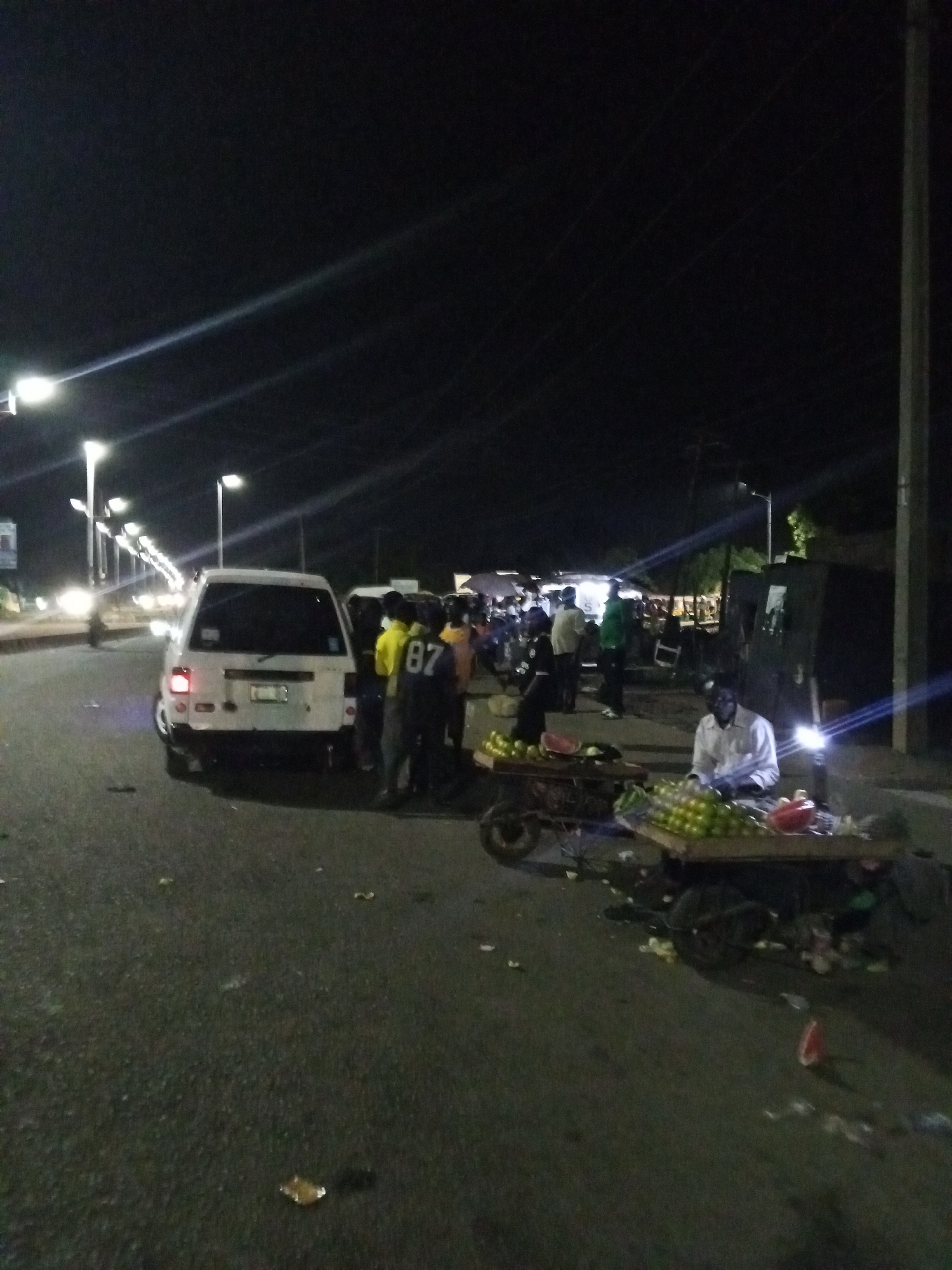 The image depicts a street scene at night in Bukuru, Nigeria. Several people are gathered near a white minivan parked on the road. A fruit vendor is seated next to a cart laden with produce, including oranges and watermelon. The street is illuminated by overhead streetlights. There are some fruits on the ground, possibly rejects. Other people appear to be milling about near the roadside.