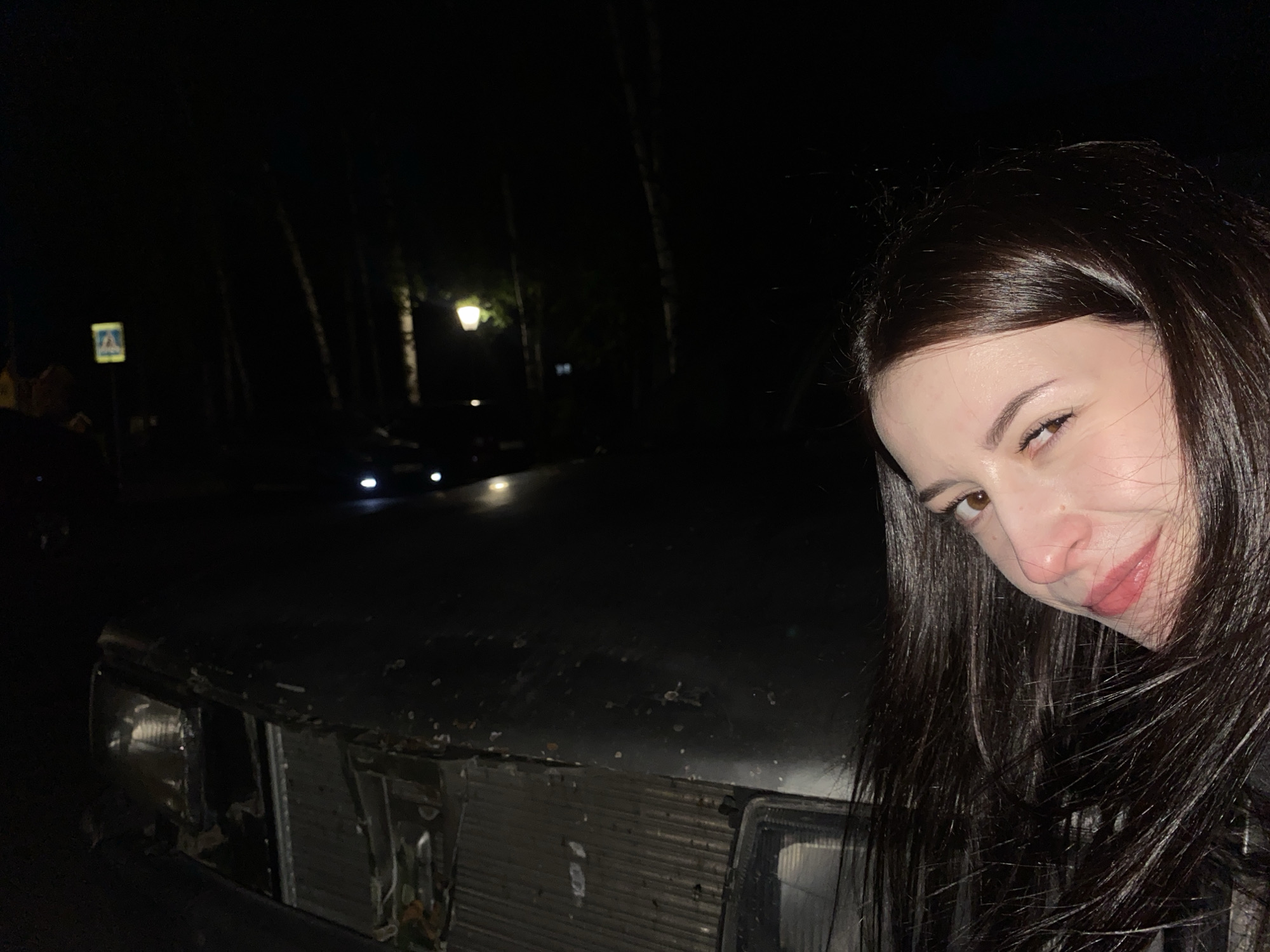 The image presents a nighttime outdoor scene, with a woman and a vehicle as the primary subjects. On the right side of the frame, a woman with long, dark hair is shown from the chest up, looking towards the camera with a slight, playful smile. Her face is partially visible, showing her brown eyes and rosy lips.
To the left, the front section of a car or similar vehicle is prominent. The vehicle appears old or damaged, with parts of its grille missing or broken, exposing internal components, and its headlights are visible.
In the background, several tall trees are silhouetted against the dark night sky. Scattered streetlights cast a warm glow, providing illumination to parts of the scene. In the far left background, a square blue sign with a white icon, resembling a pedestrian crossing sign, is faintly visible.
The overall impression is a casual, low-light photograph, possibly a selfie, taken next to a car in an outdoor setting at night, likely in an urban or suburban environment. The woman's expression conveys a sense of calm or amusement. No discernable text is readable on the visible sign or elsewhere in the image.
