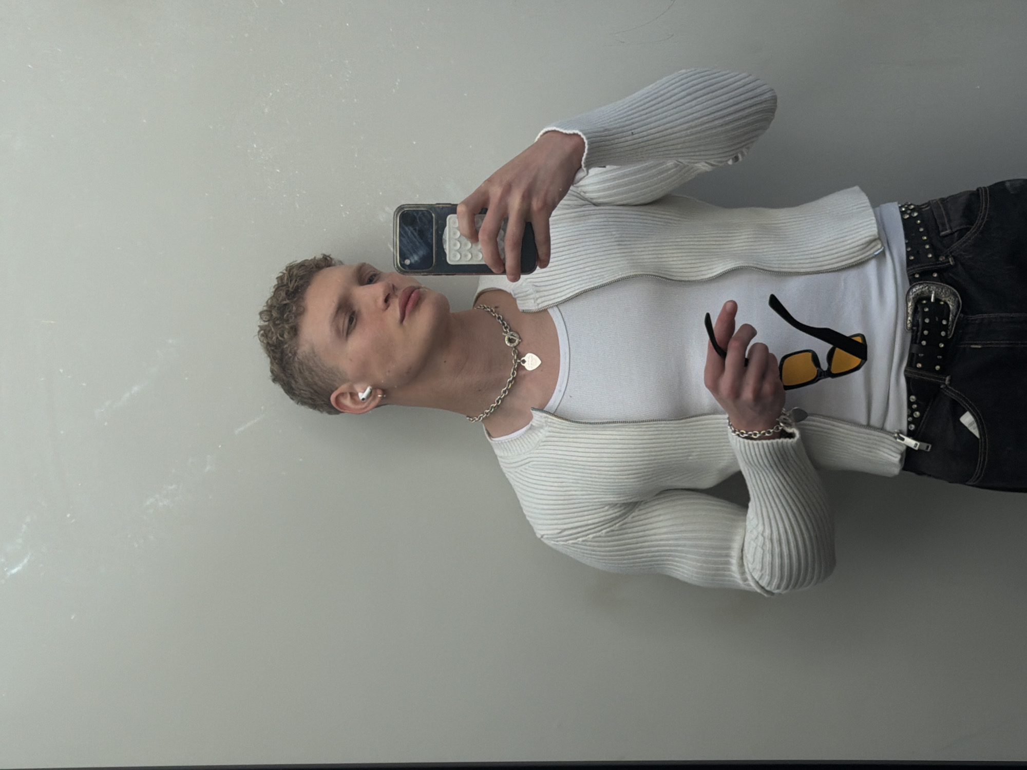 A young, light-skinned individual with short, curly blonde hair is depicted in a rotated mirror selfie, appearing to lie on their left side. They are looking towards a black smartphone, held in their right hand, which is equipped with a white pop socket or grip. The person is dressed in a white ribbed, long-sleeved zip-up cardigan over a plain white t-shirt. They wear dark jeans with a studded belt featuring a large, ornate silver buckle.
Notable accessories include a chunky silver chain necklace with two heart-shaped charms, one of which clearly displays "TIFFANY & CO." A matching silver chain bracelet adorns their right wrist, and a silver ear cuff is visible on their left ear. In their left hand, they casually hold a pair of yellow-tinted sunglasses with black frames. The background is a plain, very light-colored reflective surface, possibly a mirror or glossy wall, which shows some subtle marks or dust. The setting appears to be indoors in London, United Kingdom, conveying a relaxed and casual moment.