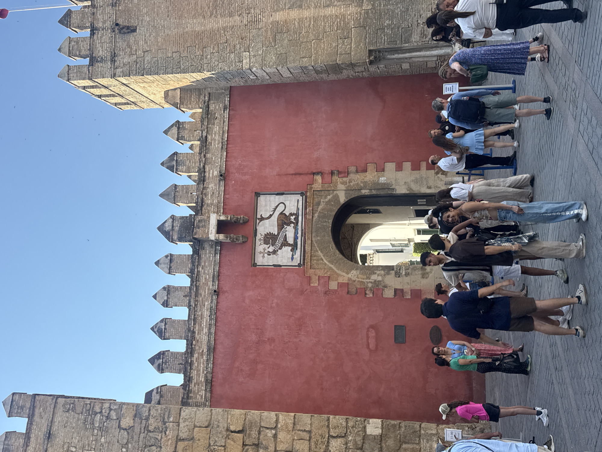 The image depicts the Puerta de la Macarena in Seville, Spain, a historic gate in the city walls. A group of tourists are gathered in front of the gate, which is a vibrant red color and adorned with a tiled crest above an arched entryway. The sun casts shadows, indicating it is either morning or late afternoon. The cobblestone ground and the stone architecture, including crenellated battlements on the towers, suggest a warm climate and a well-preserved historical site. The people are dressed casually, some with backpacks, indicating they are likely exploring the city. One person is taking a photo of the gate.