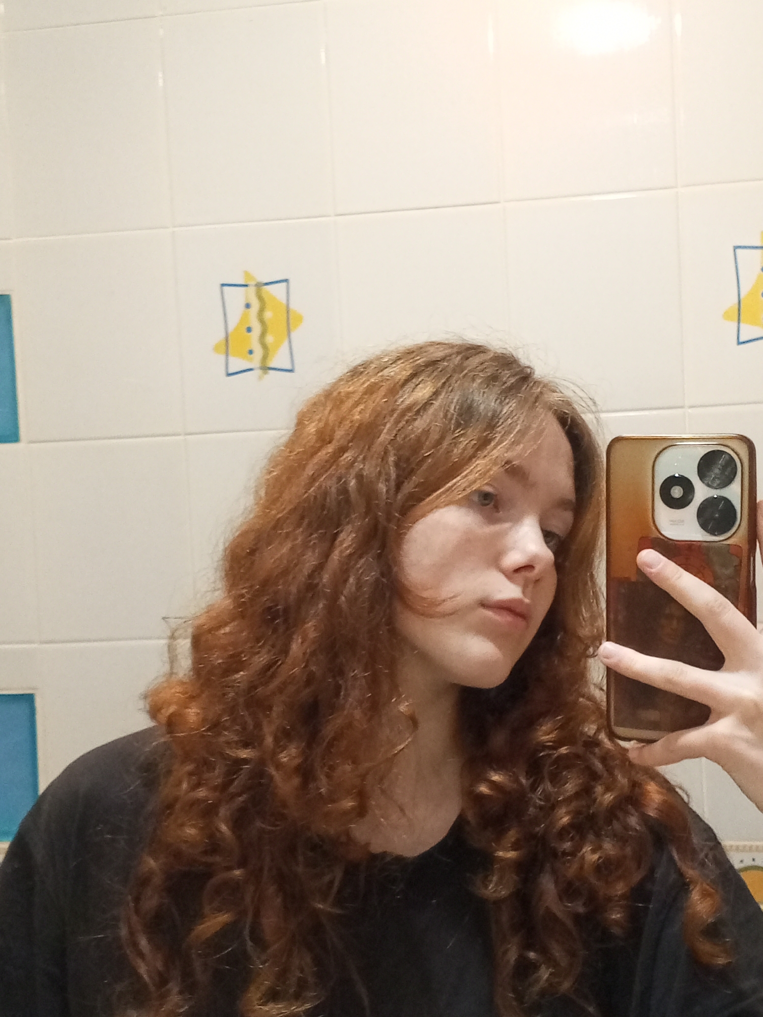 A young woman with reddish-brown curly hair takes a selfie with a gold-colored smartphone. The background is a tiled wall with white tiles and blue and yellow decorative tiles. The woman is wearing a black shirt. There is no confirmed location in the city of Chaikovsky, Russia.