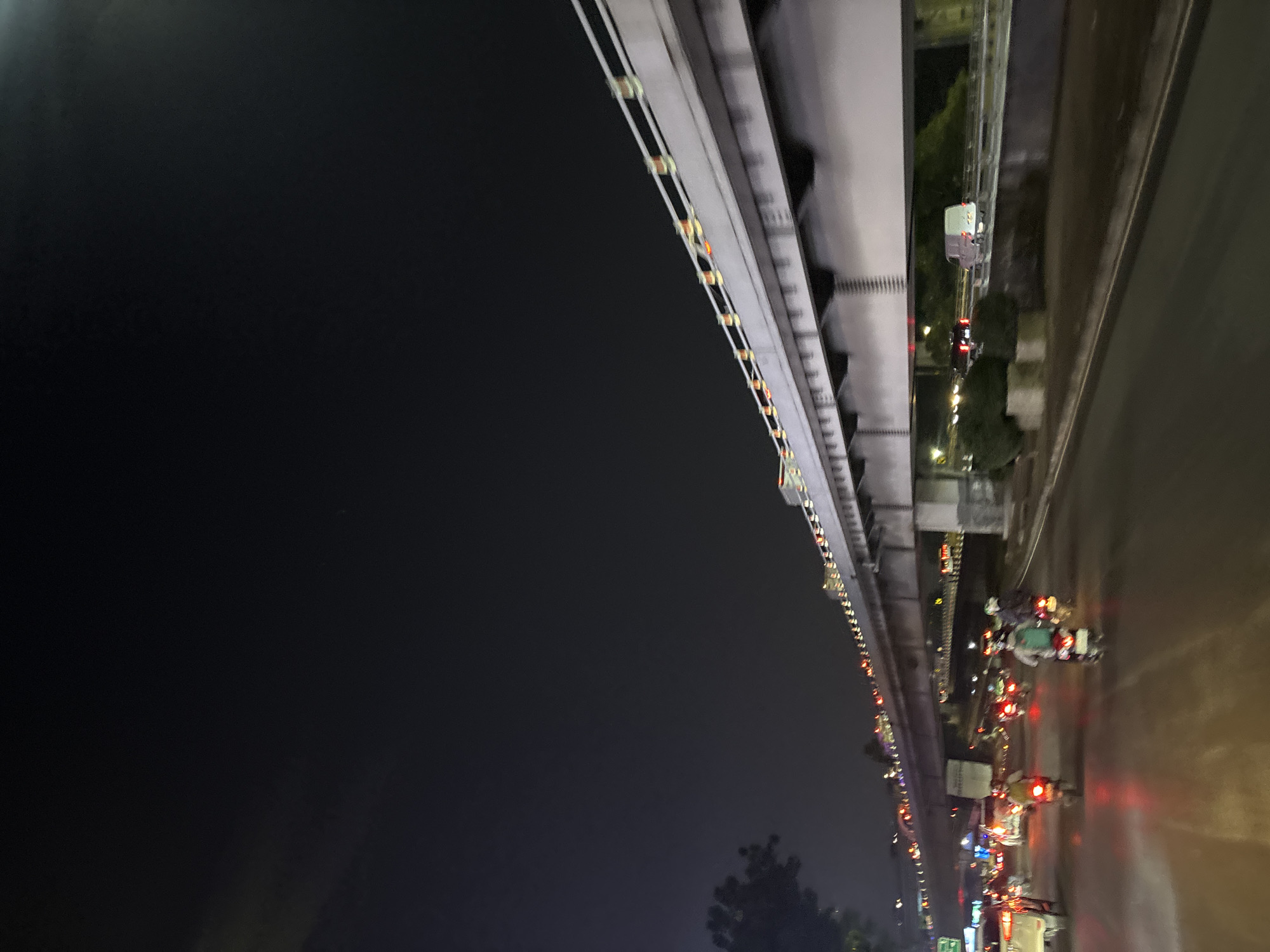 The image captures a night-time street scene in West Jakarta, Indonesia, characterized by moderate traffic and overhead infrastructure. A multi-level bridge or flyover dominates the upper portion of the frame, illuminated by strings of decorative lights along its edge, with supporting concrete pillars visible. Below the bridge, the road is wet, reflecting the headlights and taillights of vehicles. Several motorbikes are in motion, with riders visible, some wearing helmets. Other vehicles, including cars and a van, are also present on the road. The surrounding environment suggests an urban setting with some trees visible on the left side of the frame. The overall atmosphere is that of a busy city at night, with the flow of traffic continuing under the elevated structure.