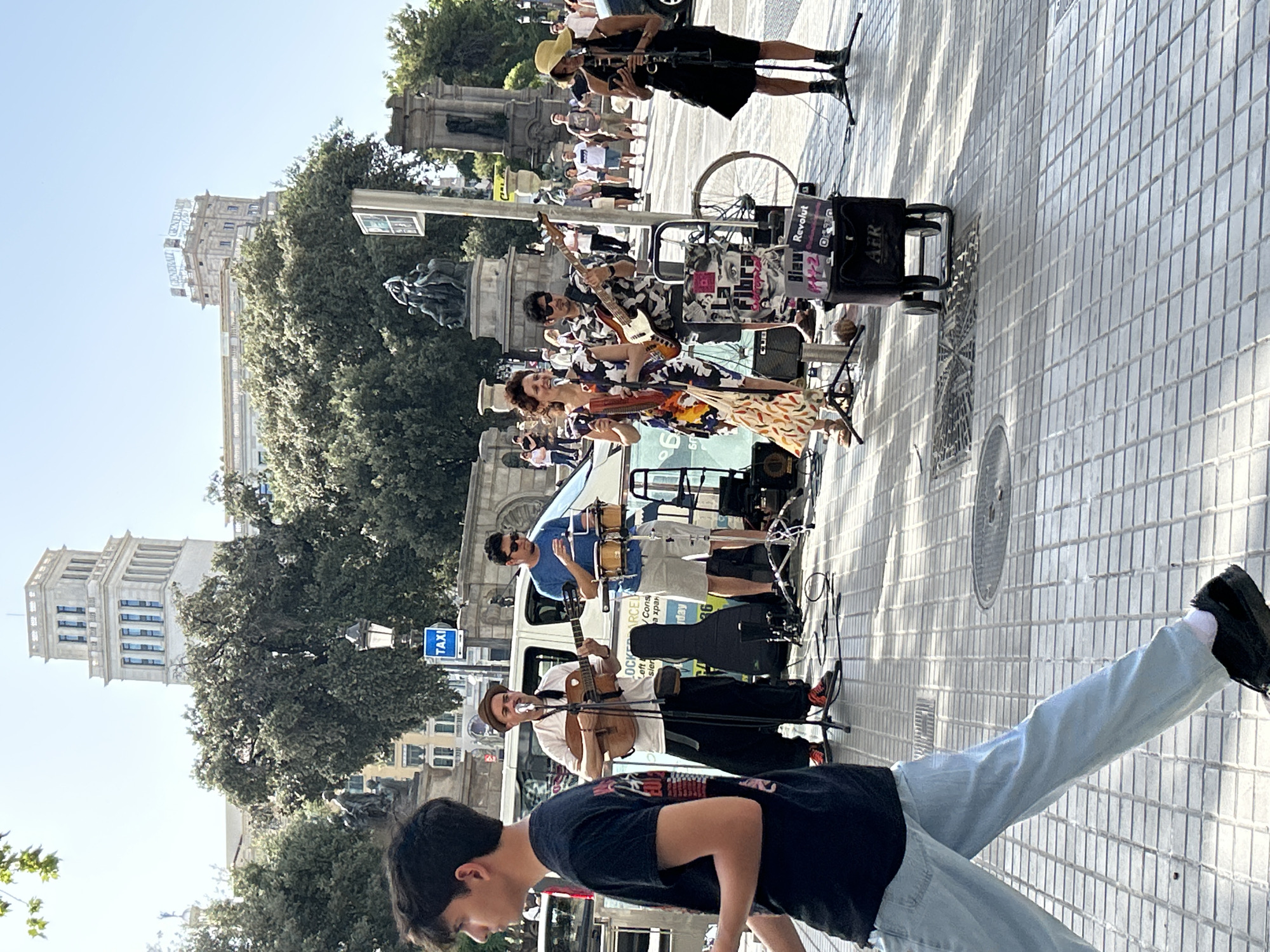 The image depicts a street scene in Barcelona, Spain, with a group of street performers playing music. In the foreground, a young man wearing a black t-shirt and light blue jeans walks past. Behind him, a band is set up on a paved area.
The band consists of four musicians. The person on the far left is wearing a hat and playing an acoustic guitar and singing into a microphone. Next to him, a person in a blue t-shirt and shorts is playing a set of bongos. To their right, a woman in a colorful dress is playing a melodica and a stringed instrument. Behind her, a person in a camouflage shirt is playing a bass guitar. A woman in a black dress and a straw hat stands on the far right, holding a phone and seemingly singing into a microphone, though her instrument is not clearly visible.
Several musical instruments and equipment are visible, including guitars, bongos, a bass guitar, microphones, amplifiers, and instrument cases. A small trolley with a speaker and what appear to be CDs or records for sale is placed in front of the performers. A bicycle wheel is also visible.
In the background, there are trees, buildings, and statues. A prominent white building with a tower stands tall in the distance, along with another building with a "GENERALI" sign. A blue "TAXI" sign is also visible. Pedestrians are walking in the background, and there are several cars parked on the street. The overall scene suggests a lively urban environment with a focus on street entertainment. The paving stones of the sidewalk are distinctive, composed of hexagonal and square tiles.