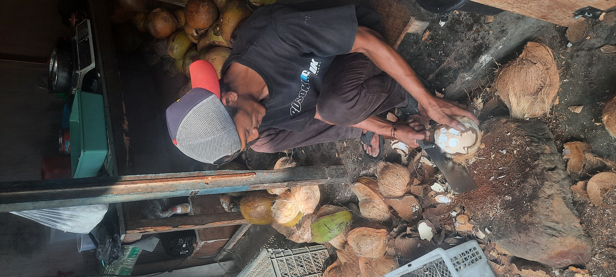 A man wearing a red and blue baseball cap and a dark t-shirt is crouched down, preparing coconuts in what appears to be an outdoor market stall. He is using a large, curved knife to open a coconut, which rests on a rough wooden chopping block. Several opened coconut husks and whole coconuts are scattered around him on the ground, which is dirt and debris. In the background, shelves are filled with various items, including what looks like a digital scale, plastic containers, and bags of produce. The overall environment suggests a rustic, open-air setting, possibly in a rural area of Selopuro, Indonesia.