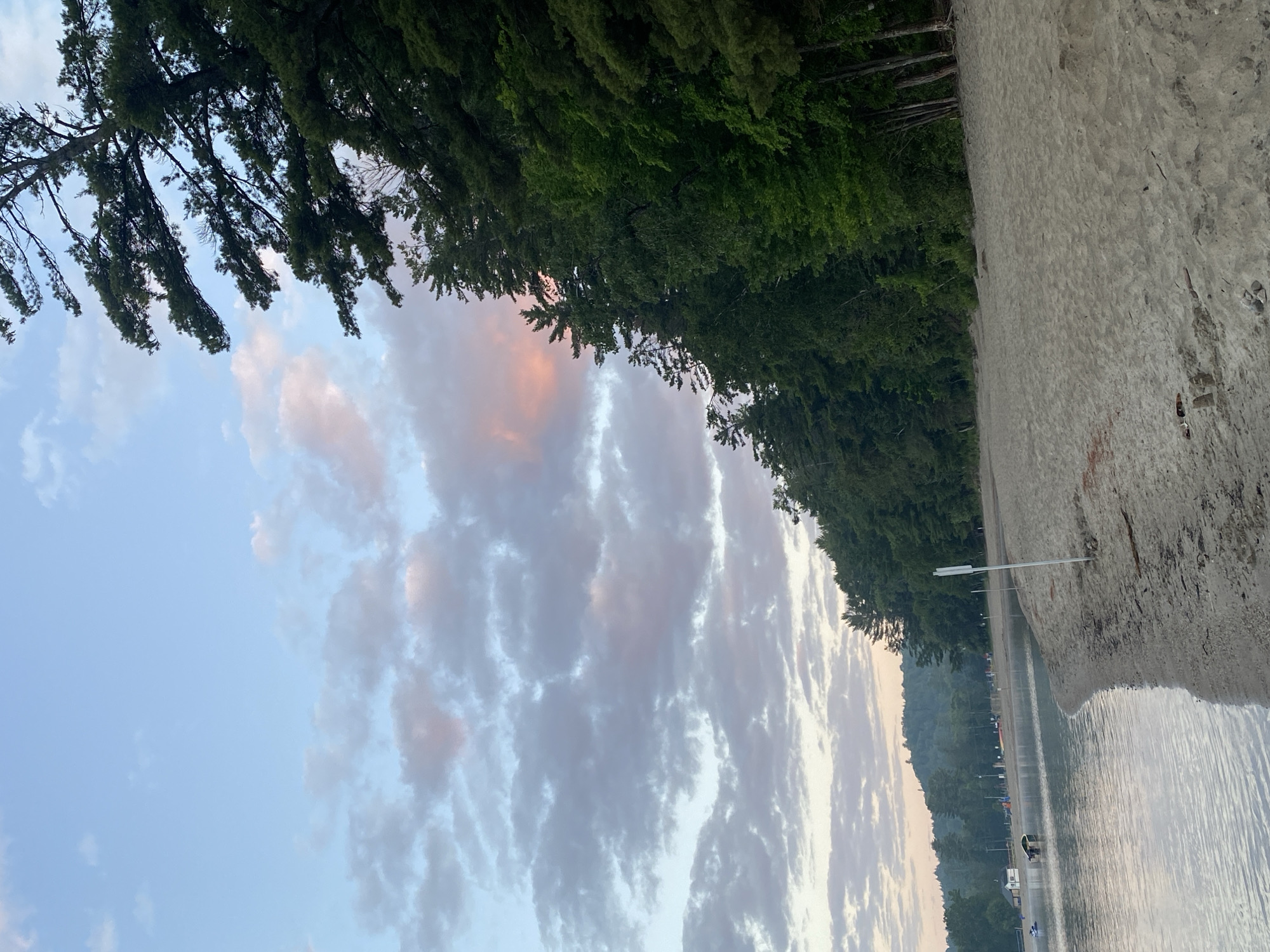 The image depicts a serene beach scene at Parc Plage Pop, Duhamel, Canada, during either sunrise or sunset. The sky is a pale blue with scattered clouds tinged with pink and orange hues. To the right, a dense line of evergreen trees forms a backdrop, with some branches extending towards the sky. The beach itself is composed of light-colored sand, with some scattered debris and footprints. Gentle waves lap at the sandy shore, and the water appears calm. In the distance, across the water, there are faint outlines of hills and more trees. There are a few people visible on the beach in the background, along with some boats and structures near the water's edge, suggesting a recreational area. The overall atmosphere is peaceful and natural.
