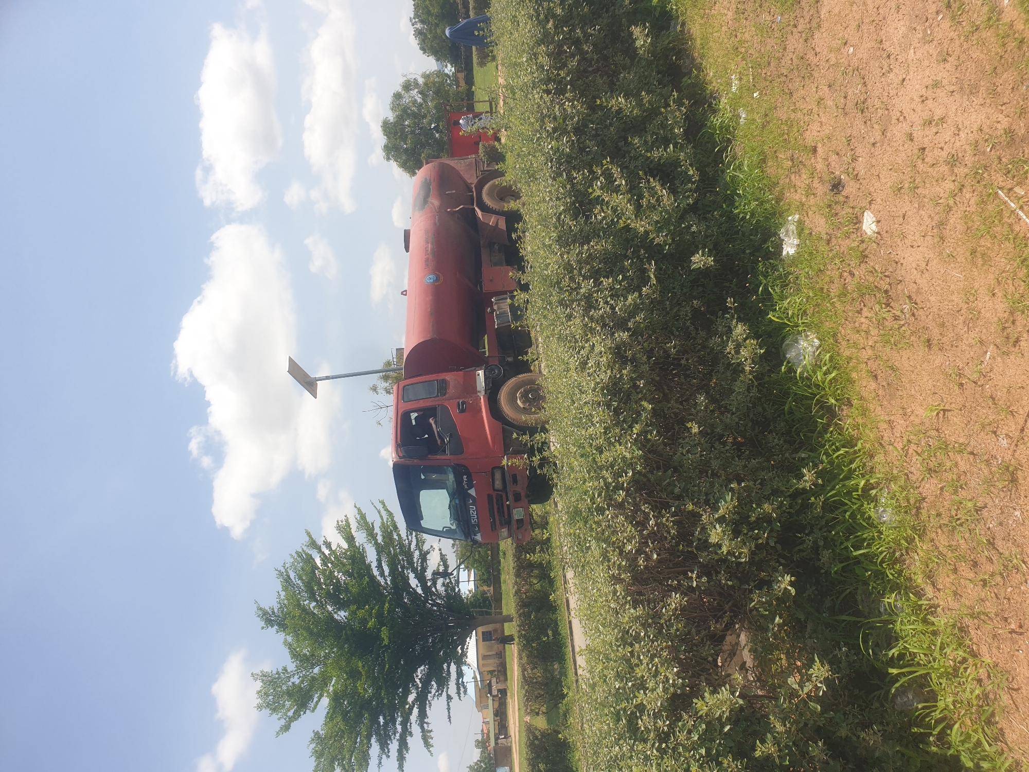A red ISUZU FTR water truck is parked on a paved road in Jalingo, Nigeria. The truck has a large red water tank mounted on its chassis. The cab of the truck is visible, and a person can be seen sitting in the driver's seat. In the background, there is a green tree, a building with a white roof, and several people walking. A solar-powered street light is visible near the truck. The scene is outdoors, with a blue sky and white clouds overhead. The foreground is dominated by a low, green hedge and a sandy, sparsely vegetated area. The overall impression is that of a sunny day in an urban or suburban setting.
