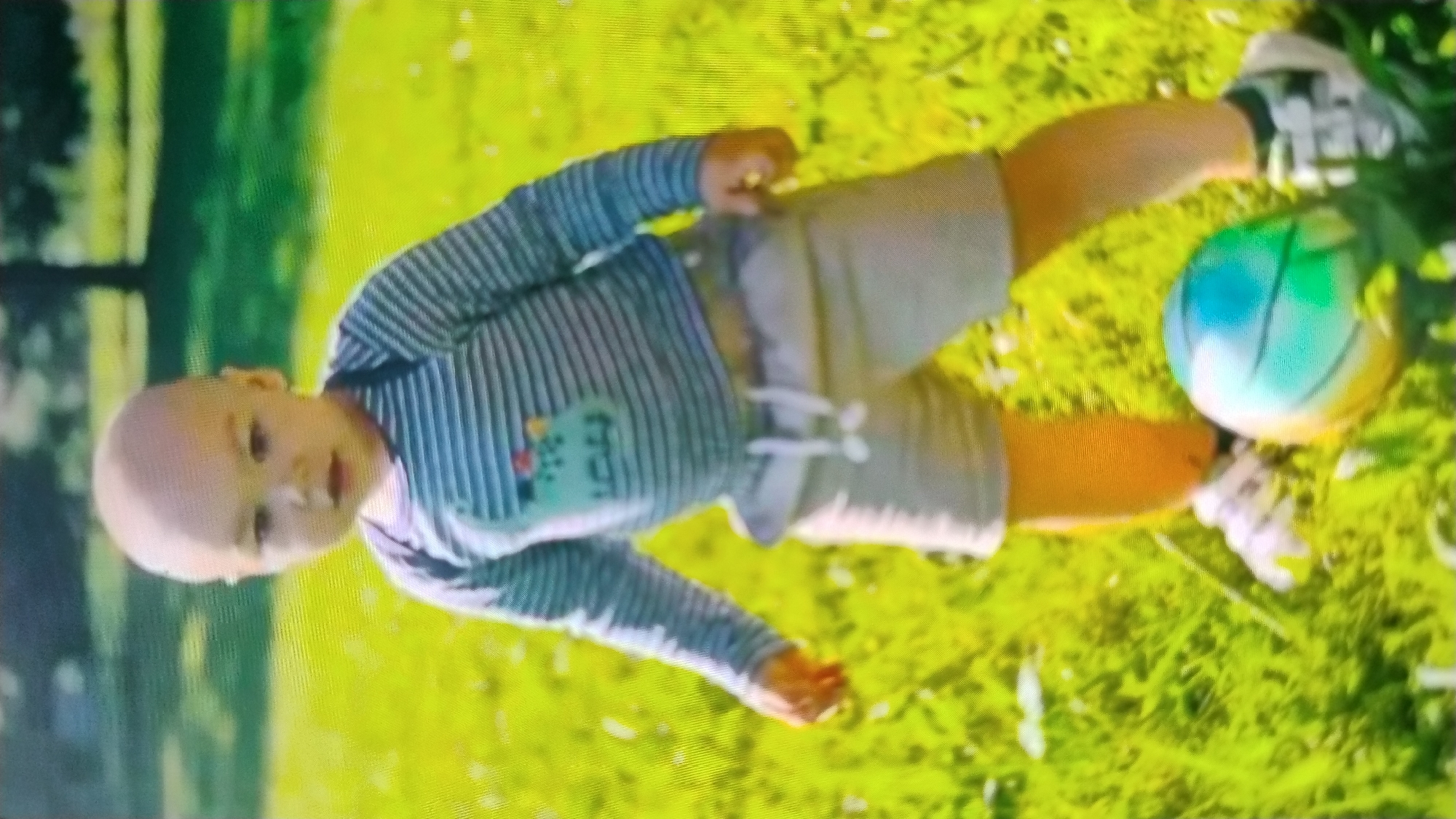 A young bald child stands in a grassy field with a soccer ball at their feet. The child is wearing a long-sleeved striped shirt and light-colored shorts. The background shows a blurred green area, possibly a park or lawn, with a tree visible in the distance. The image is taken in Mykolaiv, Ukraine.