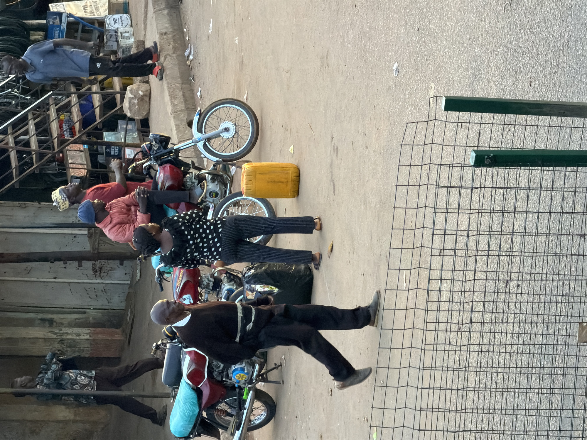 The image captures a street scene in Bukuru, Nigeria, with a focus on a bustling marketplace environment. In the foreground, a man with a dark sweater and pants walks from left to right, partially obscured by a wire fence. Behind him, two motorcycles are parked, one with a blue tarp on its seat. A yellow jerrycan sits on the ground next to the motorcycles.
In the background, several individuals are present. A man in a dark shirt and black pants stands near a set of stairs. To his left, a man in a red jacket and a blue cap is seated on a motorcycle. Another man in a red shirt and a traditional cap is standing near the stairs. A woman in a polka dot top and striped pants is standing between the two motorcycles, facing left. Further back, a man in a patterned shirt and dark pants is walking away from the camera. The environment appears to be a street with buildings and stalls lining the sides, indicative of a commercial area. The ground is paved with a rough surface.