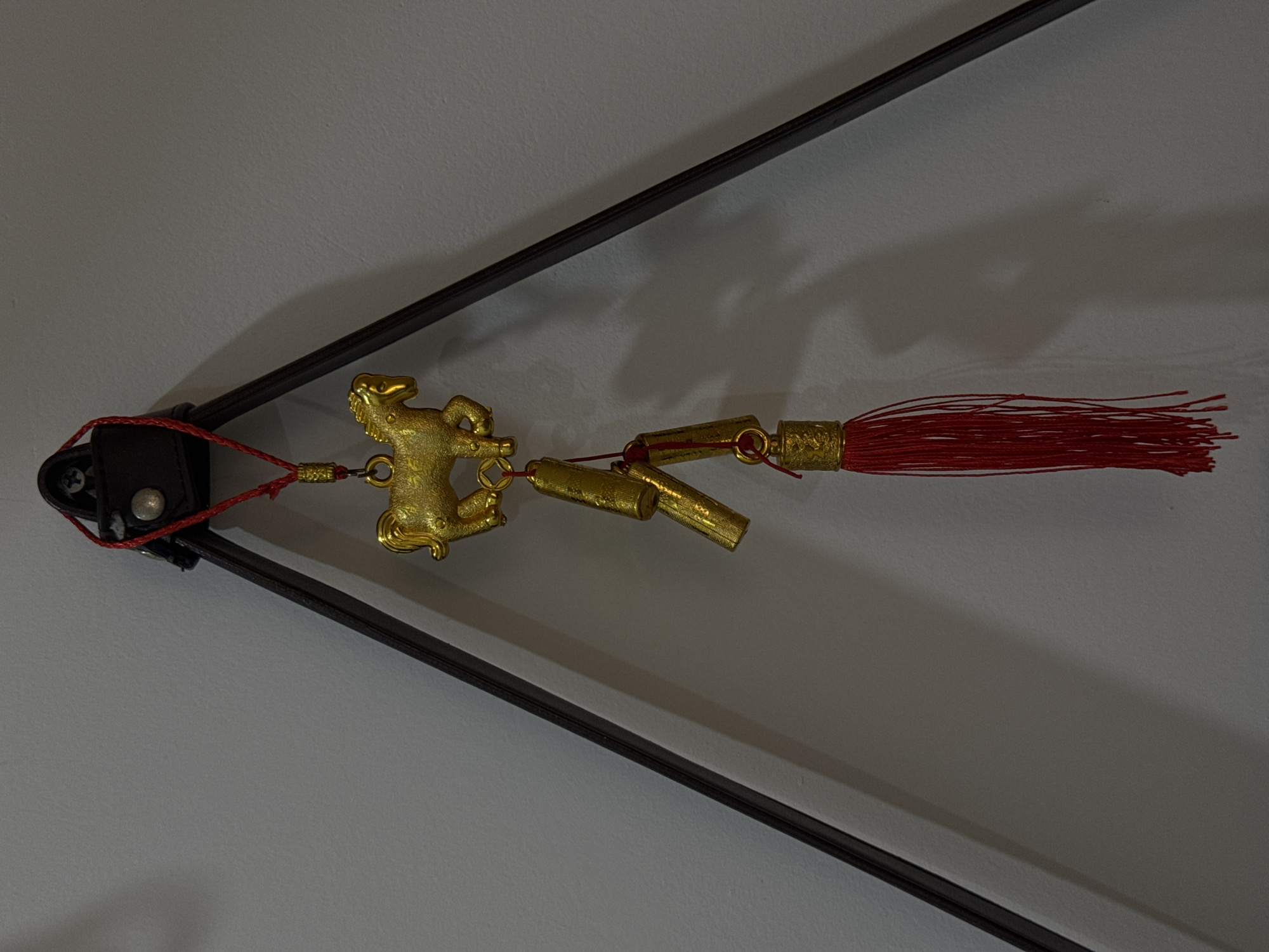 An ornamental hanging ornament, consisting of a golden horse figurine, three golden cylindrical charms resembling firecrackers, and a red tassel, is suspended from a dark triangular frame against a light grey wall. The ornament is attached to the frame by a thin red cord. The golden horse is depicted in a galloping pose. The firecracker charms have intricate etched patterns. The red tassel is full and hangs loosely. Shadows are cast on the wall by the ornament and the frame. The setting is indoors, on a wall.
