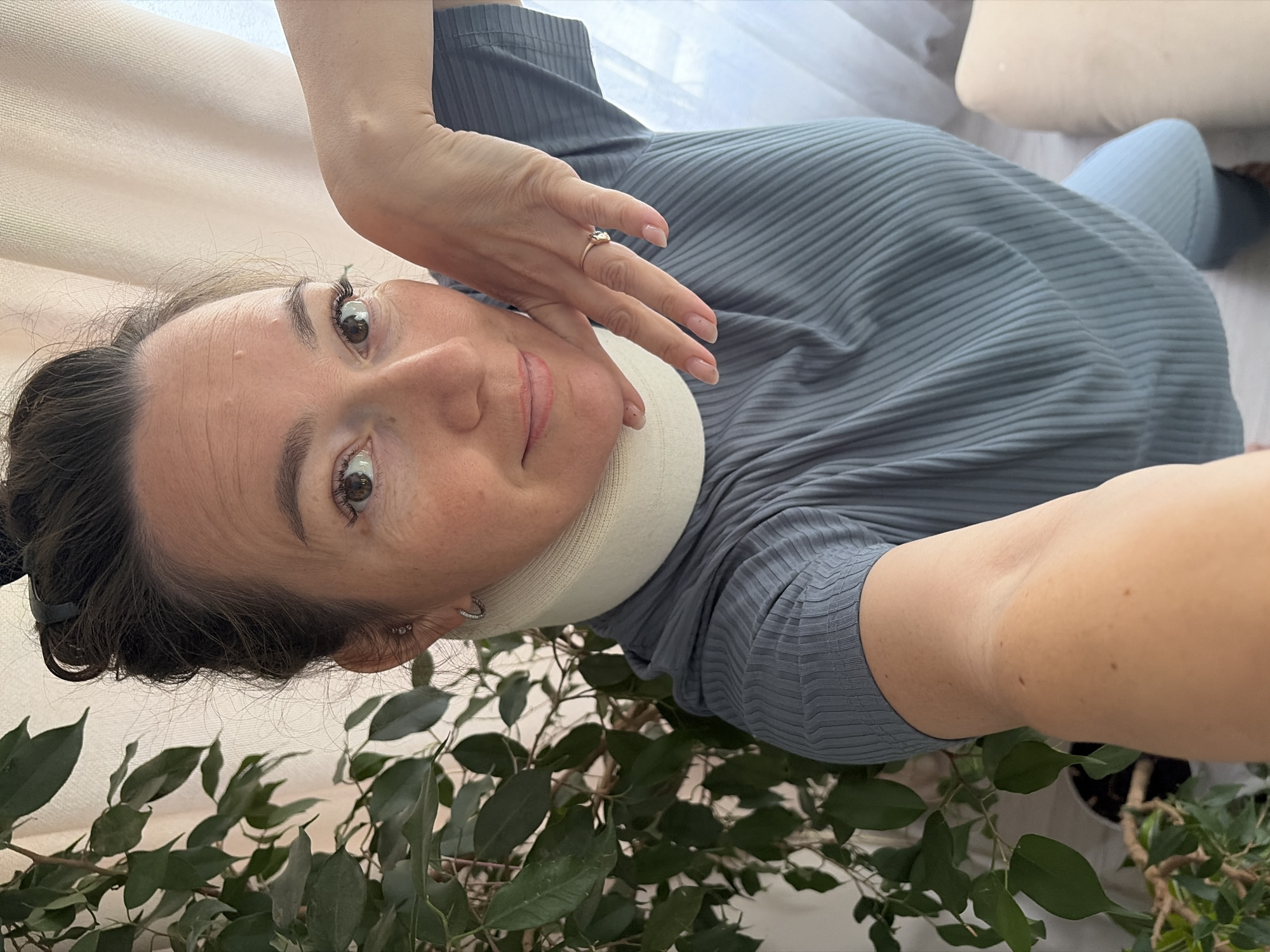 A woman is taking a selfie with a plant and a curtain in the background. She is wearing a blue ribbed shirt and a neck brace. She has her right hand on her chin and is looking at the camera. The woman has brown hair pulled up in a bun and is wearing earrings and a ring on her right hand. The scene appears to be indoors, possibly in a home. There is no indication of the city of Ольховка, Russia.