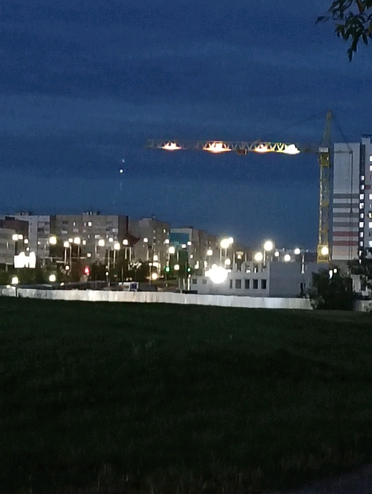 This image depicts a nighttime urban landscape in Hrodna, Belarus. In the foreground, a dark grassy area suggests a park or undeveloped land. Behind this, a line of illuminated buildings stretches across the midground, with lights visible in many windows. A construction crane, partially lit by external lights, dominates the right side of the frame, extending over the buildings. The sky is a deep blue, indicative of twilight or nighttime. The overall scene suggests a developing urban area.