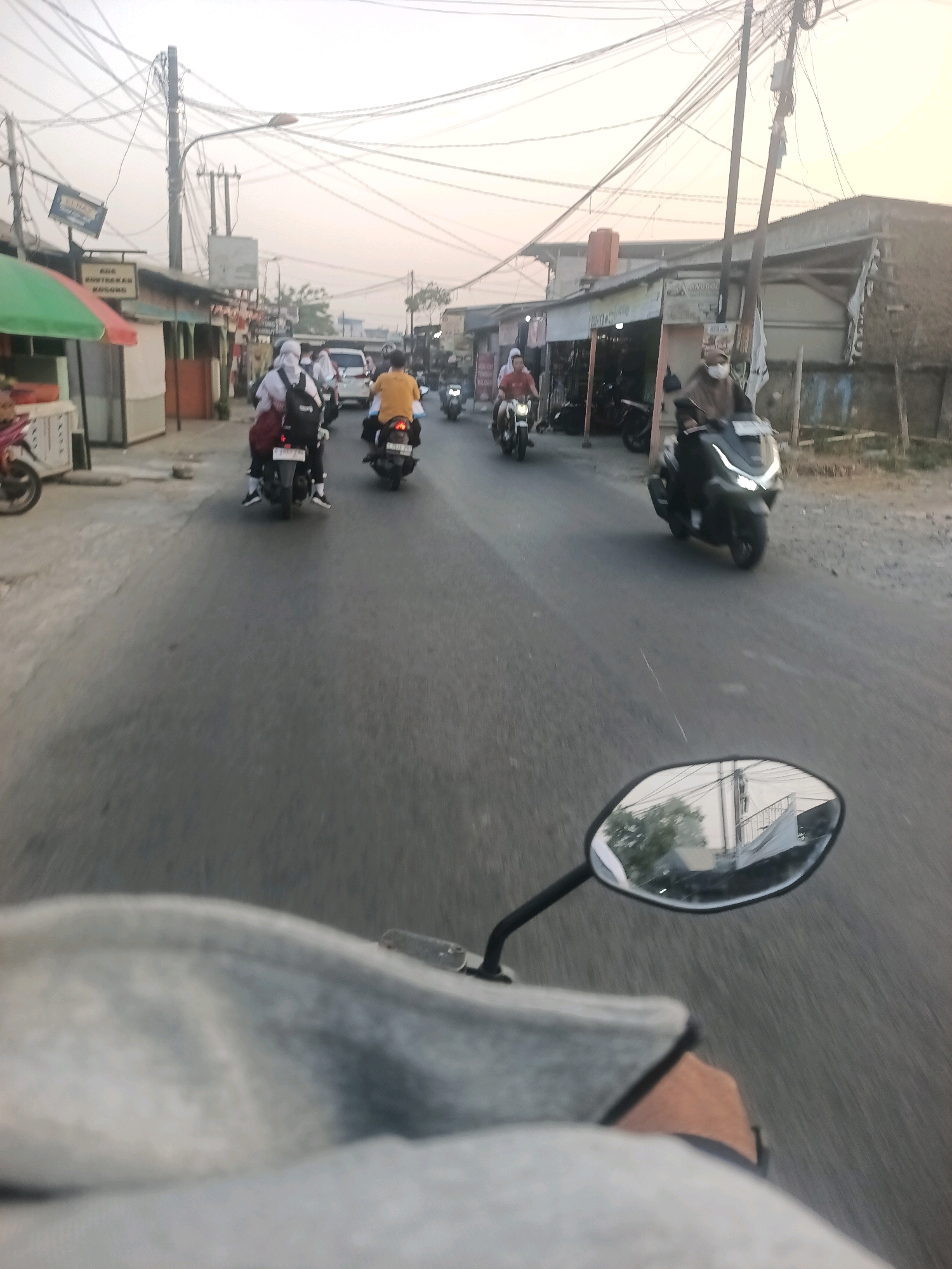 The image shows a street scene in Wanajaya, Indonesia, with several motorcycles and a white van on a paved road. The perspective is from a motorcycle rider looking forward, with the rider's handlebars and a portion of their gray jacket visible in the foreground. A motorcycle mirror is also visible on the right side of the frame, reflecting trees and buildings.
In the middle ground, a white van is traveling down the road, followed by several motorcycles. One motorcycle has two passengers wearing white headscarves and backpacks, likely students. Another motorcycle is ridden by a person in a yellow shirt. Further down the road, a dark-colored scooter is visible with its headlights on, and another person wearing a mask and headscarf is riding a motorcycle.
The street is lined with buildings on both sides. On the left, there are shops with colorful awnings, including one with a green awning. On the right, there are more utilitarian buildings and structures. Numerous electrical wires crisscross above the street, supported by poles, indicating an urban or suburban environment. The sky is overcast and a soft, diffused light suggests either early morning or late afternoon. The overall impression is a typical street scene in an Indonesian city, with everyday transportation and commerce taking place.