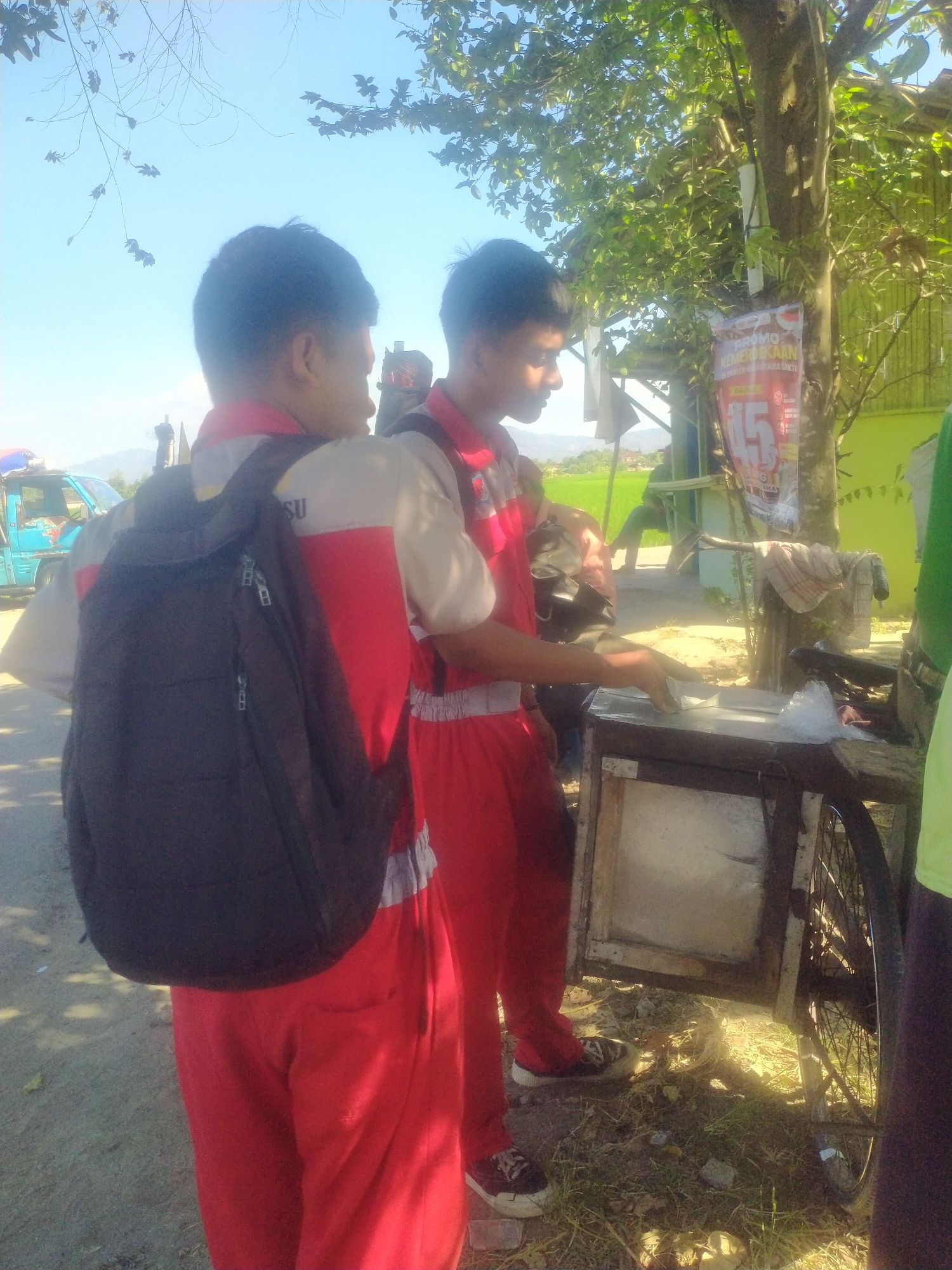 Two young males, likely students due to their matching uniforms and backpacks, are standing outdoors in a village setting, possibly in Begajah, Indonesia. The male on the left wears a red and white uniform with a black backpack, while the male on the right wears a similar red and white uniform and appears to be interacting with a food cart. The cart has a wooden frame and a metal interior, possibly for storing food. A banner is visible on a tree behind the cart, displaying Indonesian text and a numerical promotion. The environment includes vegetation, a bright blue sky, and the sun casting shadows. In the background, a light blue truck is parked. The ground is a mix of dirt and grass.