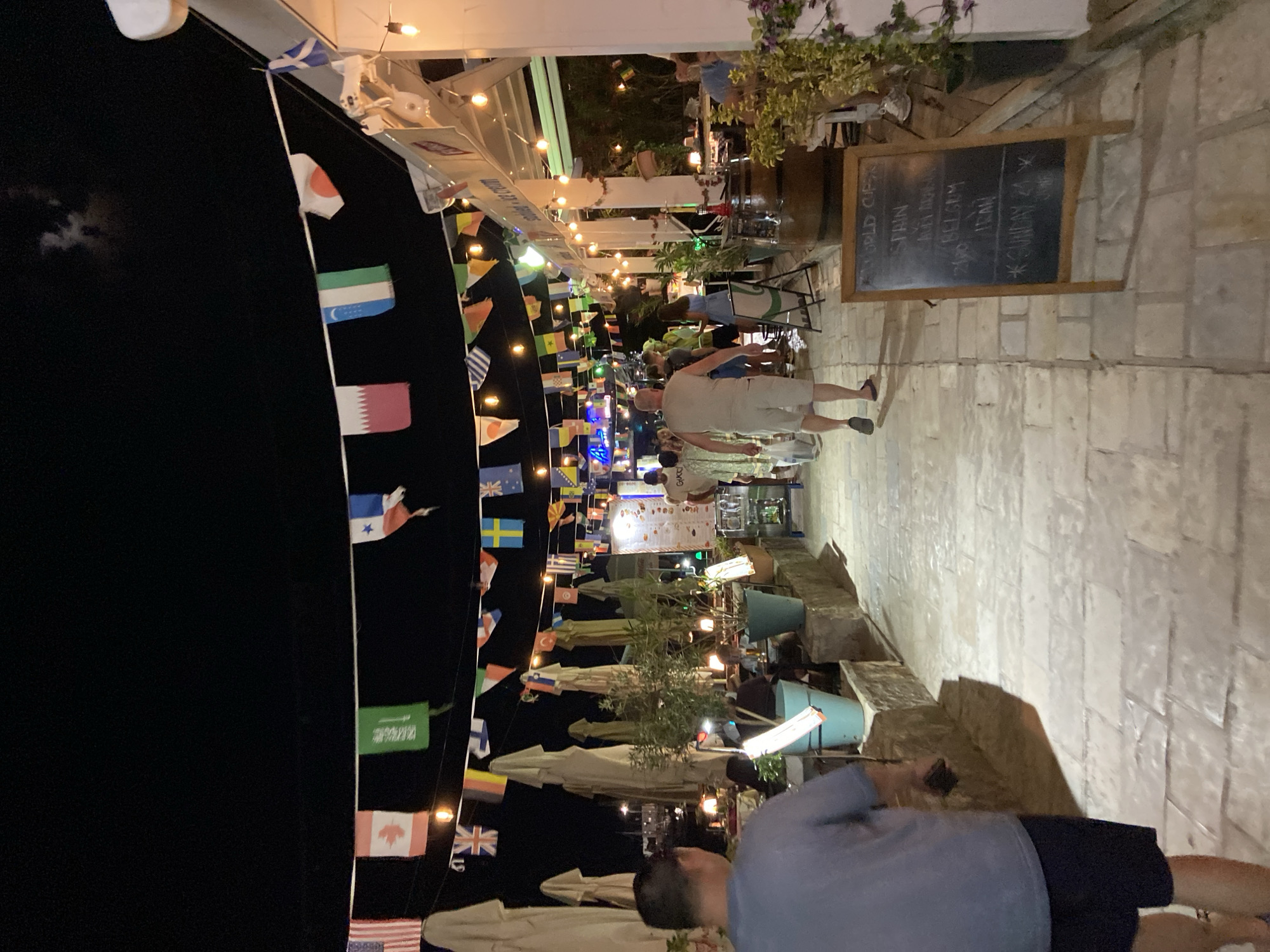 The image depicts an outdoor dining area at night in Budva, Montenegro, decorated with numerous national flags strung across the scene. A paved walkway leads through the area, lined with tables and chairs, some of which are occupied by diners. Umbrellas provide shade over some of the seating areas. String lights illuminate the scene, creating a warm ambiance. In the foreground, a man in a grey t-shirt and dark shorts walks away from the camera, holding a phone. Further down the walkway, other individuals are seen walking and dining. A chalkboard menu displays information, including "WORLD CUPS," "SPAIN," "IRAN," and "BELGIUM," along with a note about "SUNDAY." The overall impression is of a lively evening at a restaurant or bar.