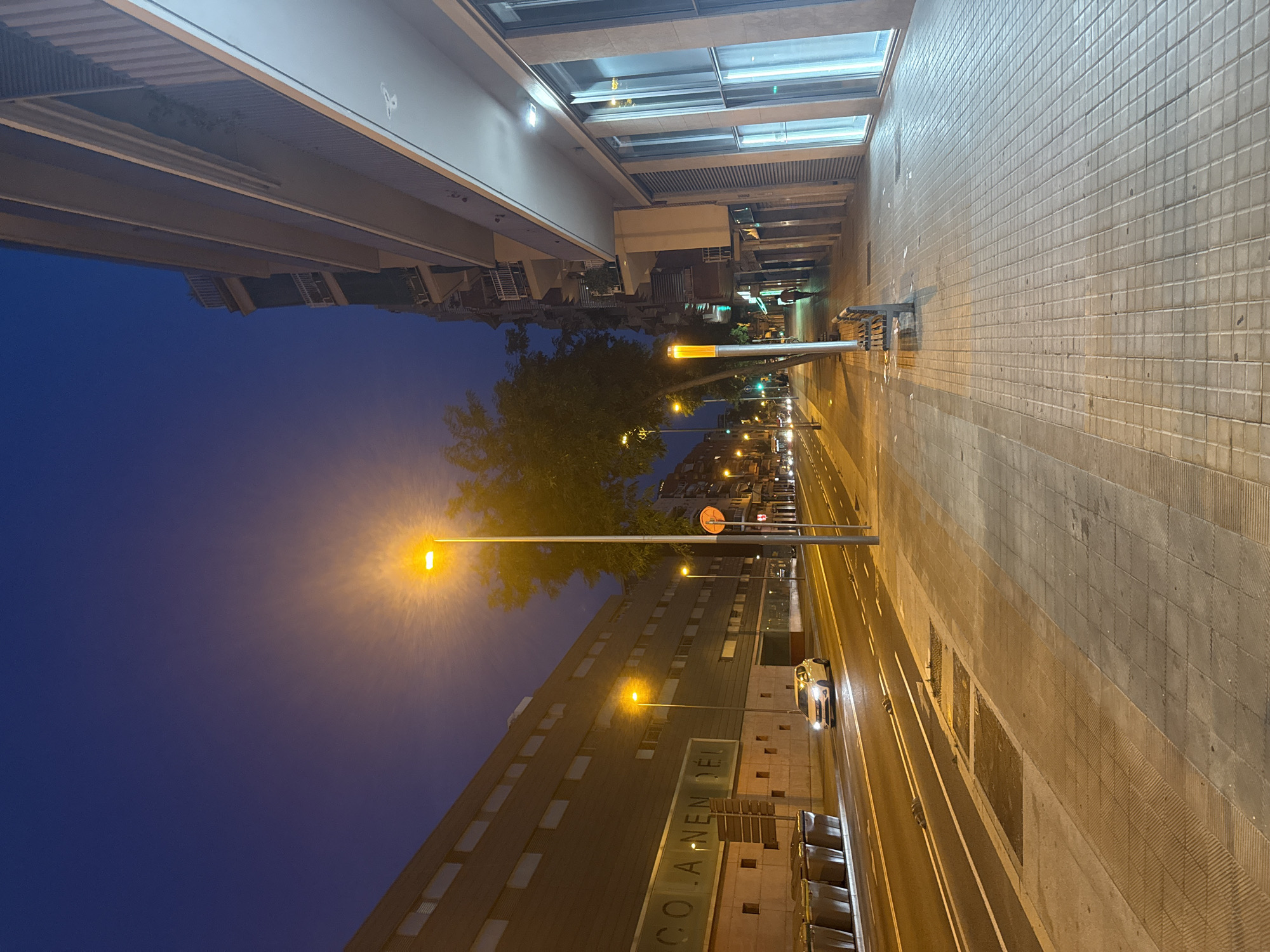 The image depicts a street scene in Barcelona, Spain, at dusk. A modern, dark-colored building with multiple floors and balconies lines the right side of the street. The building features large glass windows on the ground floor. Across the street, another large building with a dark facade and rows of rectangular windows is visible. The words "COLA NEN DEL" are faintly visible on this building's facade. A white car is driving on the road, which has lane markings and is illuminated by streetlights. Several large, dark-colored trash bins are lined up along the left side of the road. The sidewalk is paved with square tiles and has a bench. A single person is walking on the sidewalk in the distance. The sky is a deep blue, indicating twilight or nightfall.