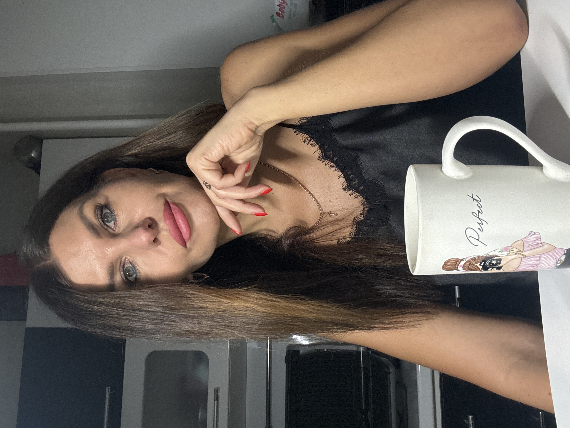 A woman with long, dark brown hair and blue eyes is sitting at a table, holding a white mug with a drawing of a woman on it that says "Perfect." She is wearing a black camisole with lace trim. Her right hand is resting on her chin, and her left arm is bent. In the background, there are kitchen cabinets and appliances. The scene is indoors, and the lighting suggests it is daytime. There is no indication of the location being in Chaikovsky, Russia.