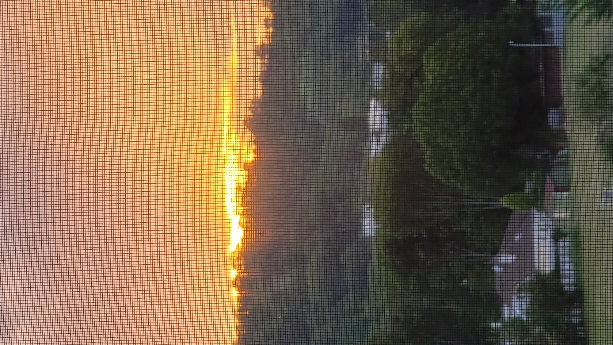 The image depicts a sunset viewed through a window screen. The sky is a vibrant orange and yellow, with the sun just dipping below the horizon, casting a warm glow over the landscape. Below the sky, a dense line of trees forms a dark silhouette. In the foreground, a residential area with houses and lush greenery is visible. The scene appears to be a suburban or semi-rural setting. The image does not provide any specific details that would allow for identification of the location within the city of Prešov, Slovakia.