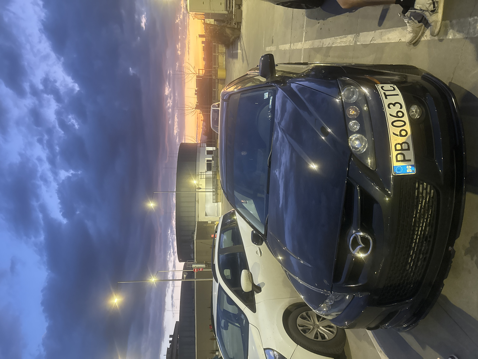 The image shows a dark blue Mazda car parked in what appears to be a parking lot in Sofia, Bulgaria, indicated by the "PB 6063 TC" license plate. Next to it is a white car. The sky is a mix of dark clouds and a bright sunset. Street lights are visible in the background. The foreground has a person wearing sneakers.