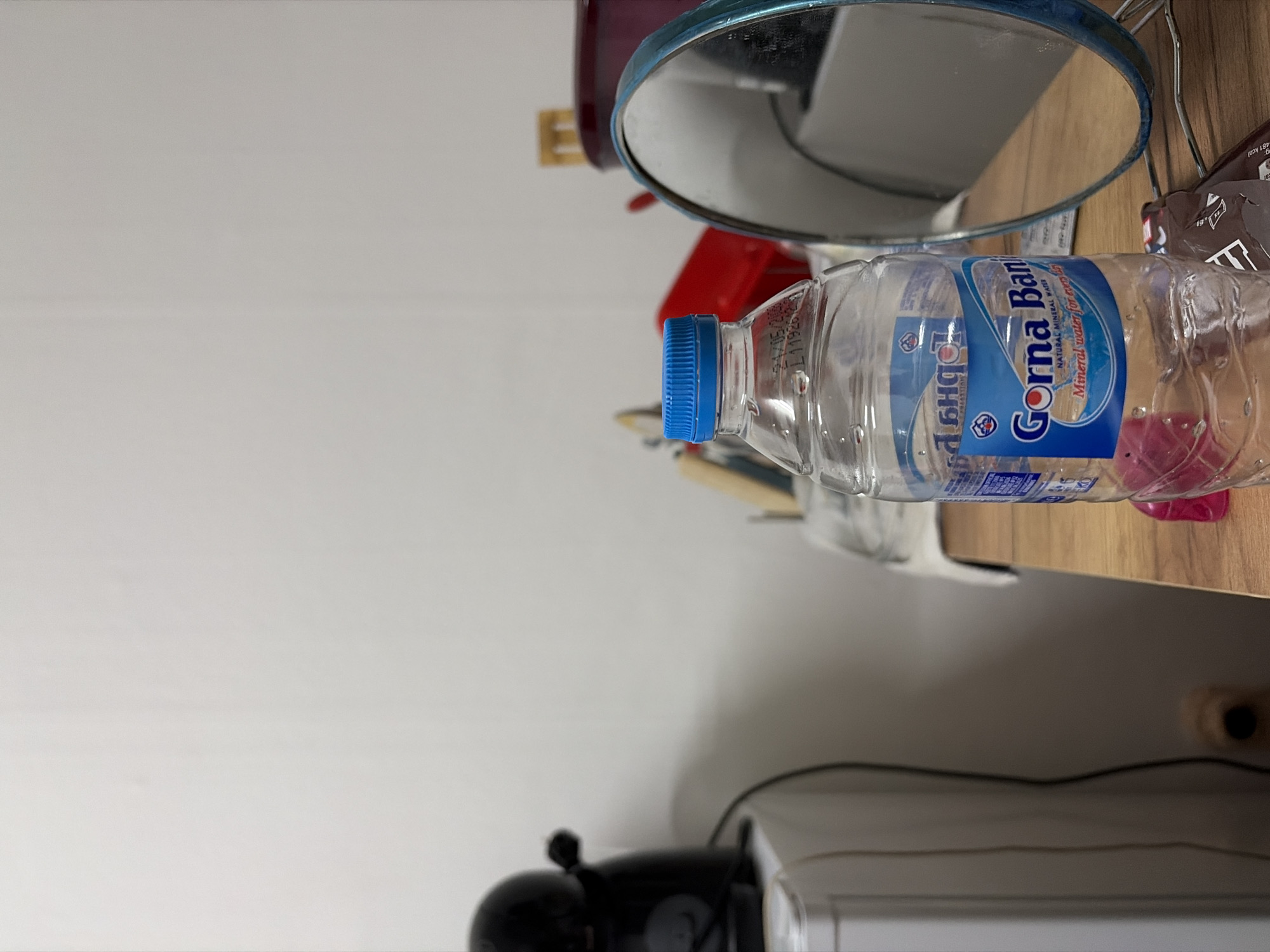 A close-up, slightly rotated image shows a cluttered shelf. A bottle of "Gorna Banja" mineral water with a blue cap and label is centrally positioned, partly obscuring other objects. A round mirror with a blue frame and a red object are visible behind the bottle. A dark-colored product, possibly chocolate, sits next to the bottle. The shelf is light brown. Below the shelf, there is a black espresso machine. The setting appears to be a domestic or personal space.