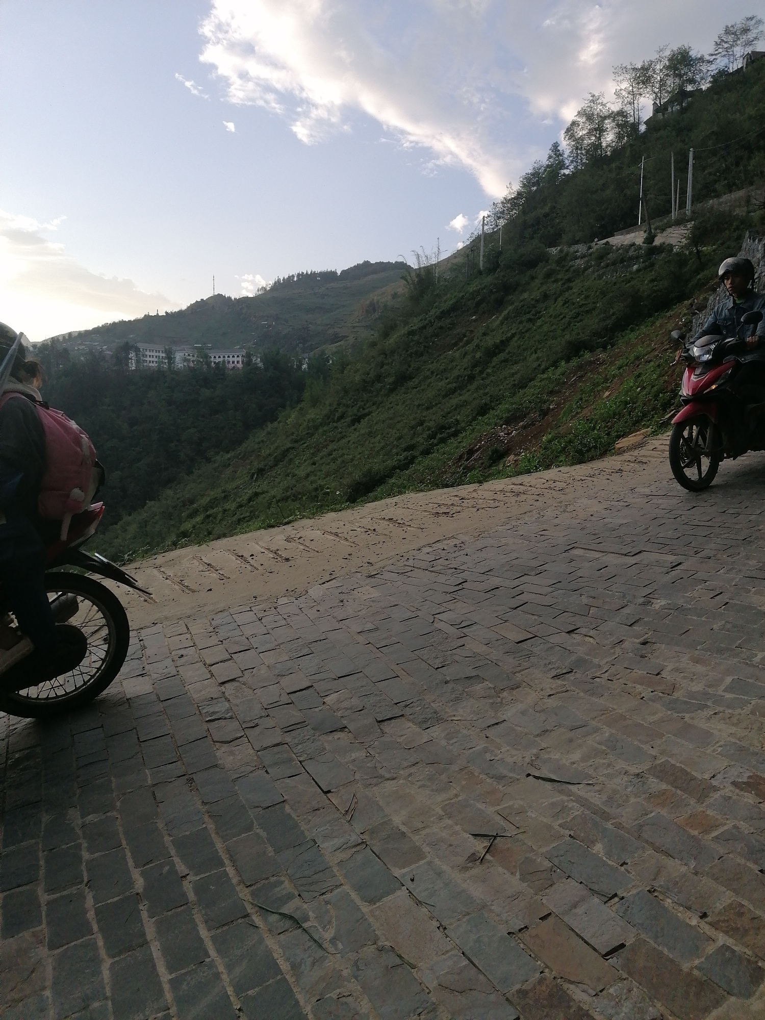 The image captures a picturesque scene from a paved road on a hillside in Sa Pa Ward, Vietnam. In the foreground, two motorbikes are visible. On the left, the rear wheel and fender of a dark motorbike are seen, with a person wearing a pink backpack partially obscured. On the right, a person in a helmet and dark jacket is seated on a red scooter-style motorbike, facing away from the viewer.
The road is distinctively paved with irregular, rectangular stone slabs, giving it a rugged, natural appearance. To the right of the road, a steep, richly vegetated hillside rises, covered in lush green trees and shrubs. Several utility poles are interspersed along this slope.
In the mid-ground and background, the landscape features undulating green mountains and forested hills. A small settlement or part of a town is visible on a distant hillside, characterized by several multi-story buildings with light-colored, possibly white or pinkish, facades. The sky is partly cloudy, with soft white clouds against a lighter blue, suggesting the warm light of either an early morning or late afternoon. The scene conveys a sense of travel through a mountainous, verdant region. There is no visible text in the image.