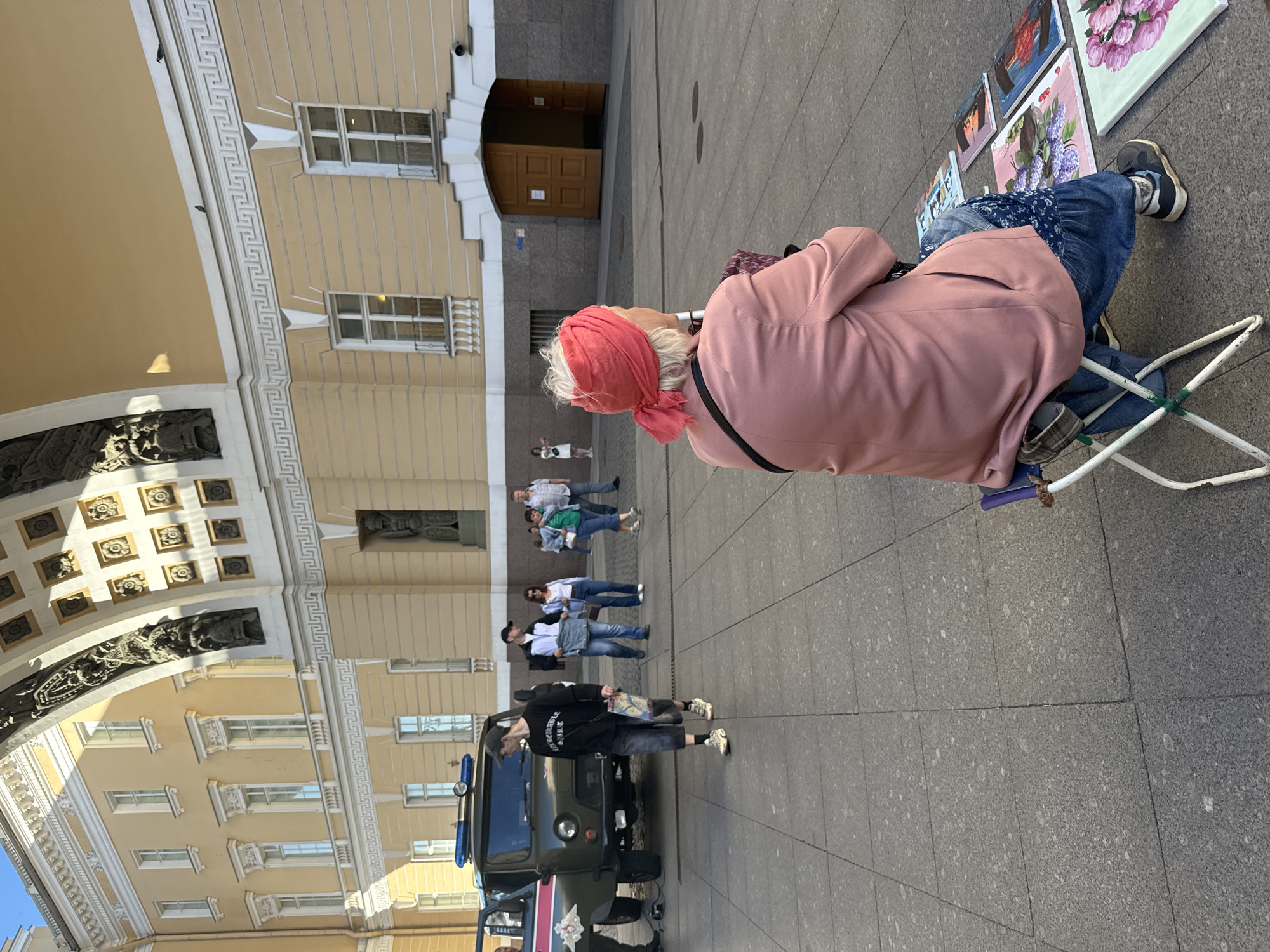 In Saint Petersburg, Russia, an elderly woman with white hair and a pink headscarf sits on a folding chair facing away from the camera. She wears a pink jacket and blue jeans, and a black bag is slung over her shoulder. Paintings of flowers and portraits are laid out on the ground in front of her. The scene takes place on a paved plaza in front of a large, ornate yellow building with classical architectural elements, under a bright sky. Several people walk in the background, and a military-style vehicle is parked to the left of the frame.