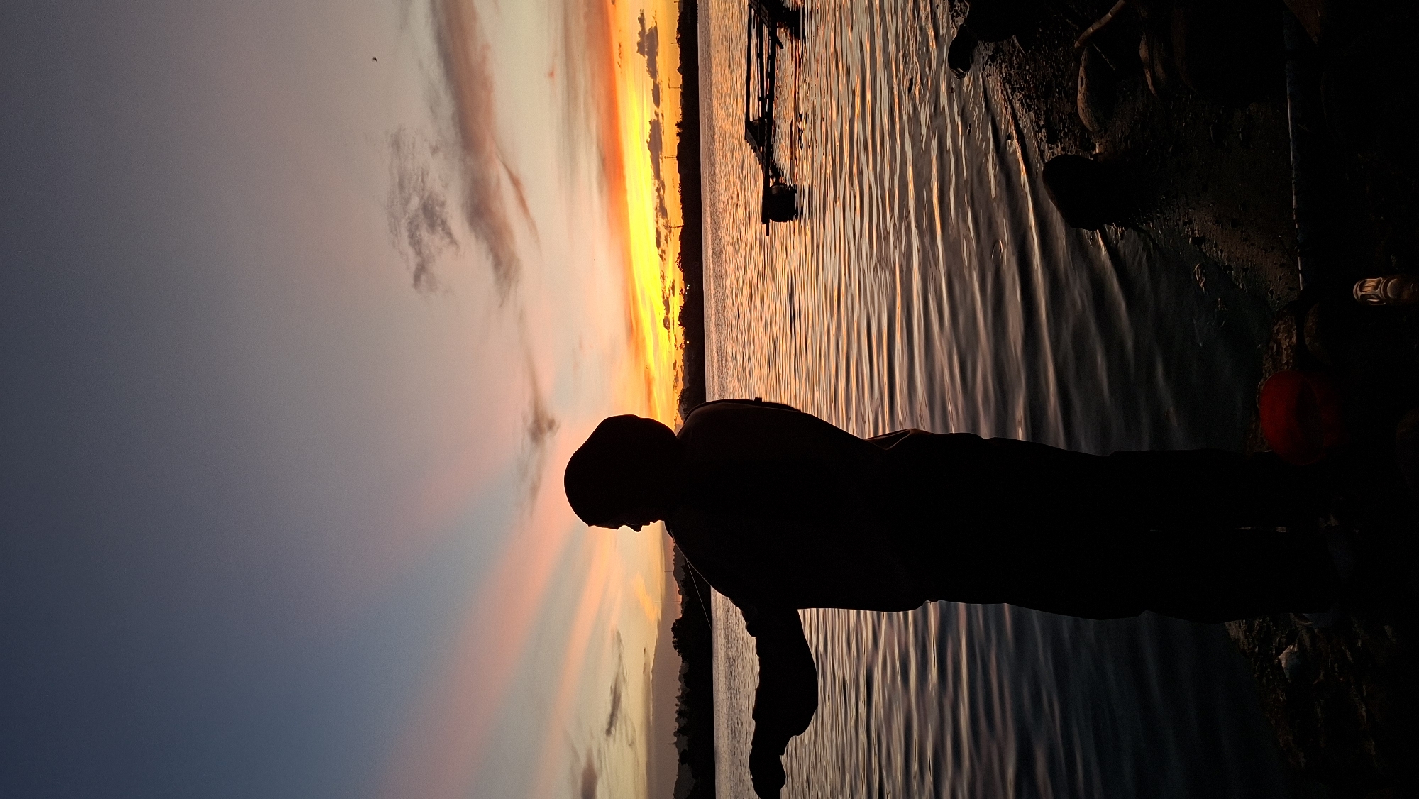The image depicts a silhouetted individual by the water during a sunset. The sky is filled with vibrant colors, ranging from deep blues and purples to fiery oranges and yellows, indicating a dramatic twilight. Wispy clouds are scattered across the sky, catching the last rays of sunlight. The water reflects the colors of the sunset, creating shimmering patterns. In the foreground, the silhouette of a person is visible, with their head bowed and facing the water, suggesting a moment of contemplation or relaxation. The person appears to be wearing a hooded sweatshirt. The setting appears to be a shoreline or waterfront, with some debris or natural elements visible along the edge of the water. The time of day is clearly sunset. The overall mood is peaceful and serene, amplified by the dramatic colors of the sky and the stillness of the water.