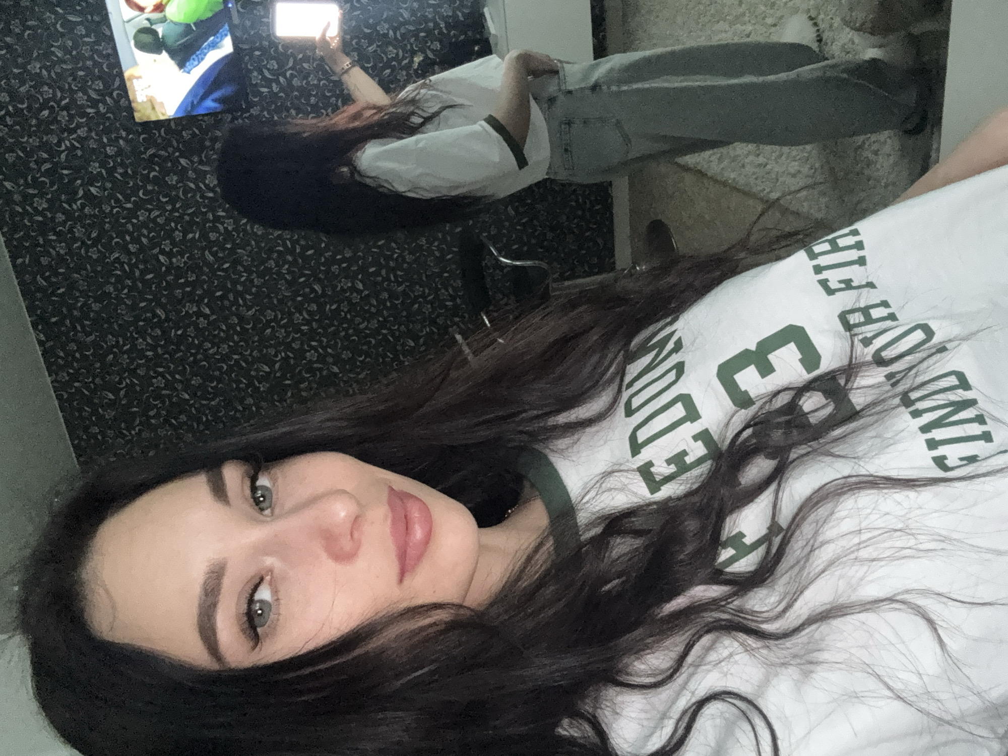 A selfie photograph shows a person with dark hair, light skin, and light-colored eyes, wearing a white t-shirt with green lettering. The person is indoors. Behind the person are another person with dark hair holding a phone and wearing a white shirt and jeans, a television, a chair, patterned wallpaper, and a carpet. The location is Chaikovsky, Russia.