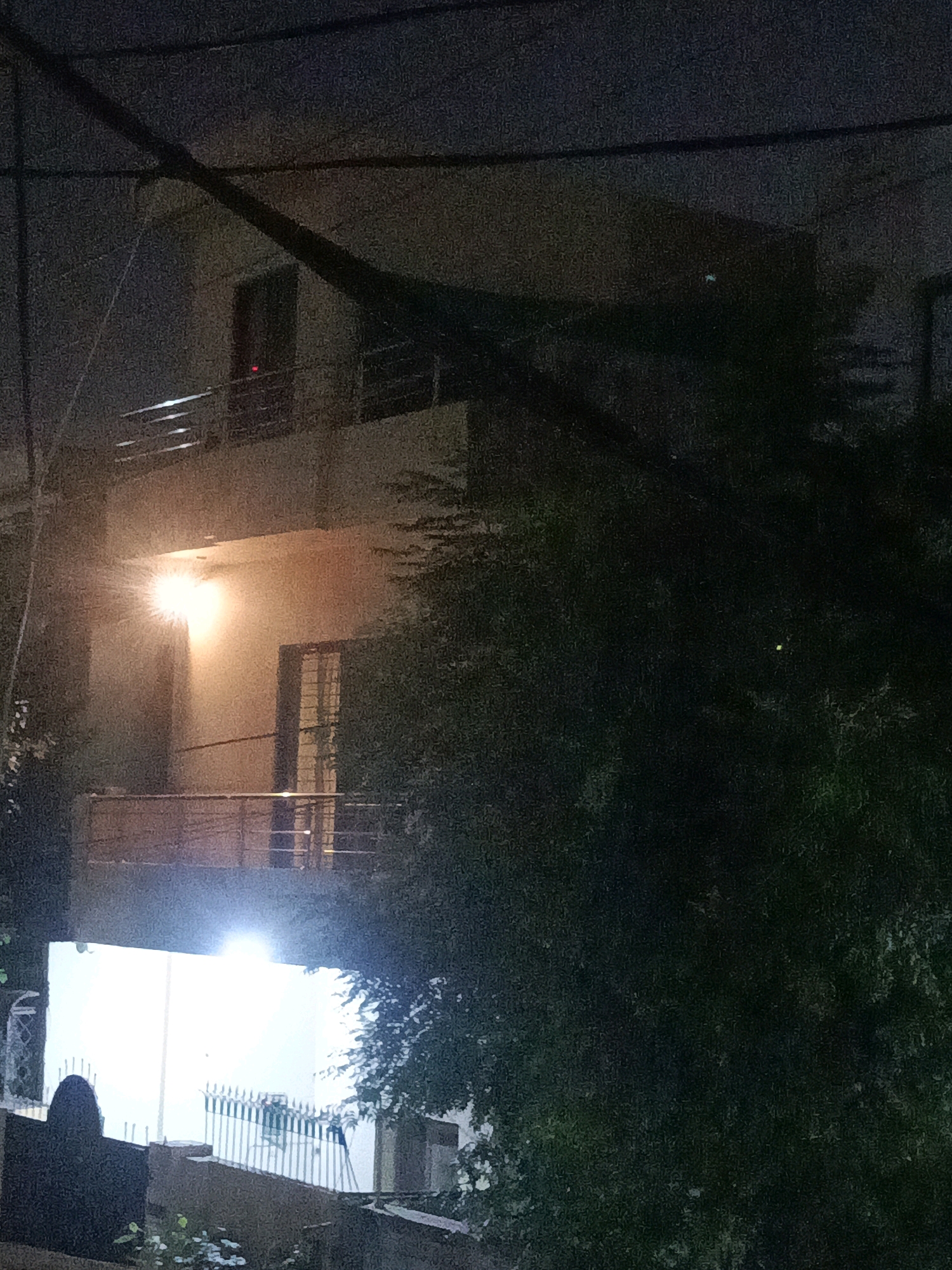 The image captures a dimly lit exterior scene, likely at night, in Rasoolpur, Pakistan. A multi-story building dominates the frame, with balconies visible on at least two levels. A bright light source, possibly a street lamp or security light, is illuminating the side of the building, casting a warm glow. Electrical wires are visible crisscrossing the sky above the building. Dense foliage, likely trees and shrubs, partially obscures the structure and the foreground. In the lower portion of the image, a white wall is brightly lit by another unseen light source, with a metal fence or railing in front of it. The overall impression is one of a residential or commercial area at night, with artificial lighting attempting to combat the darkness. No individuals are present in the image.