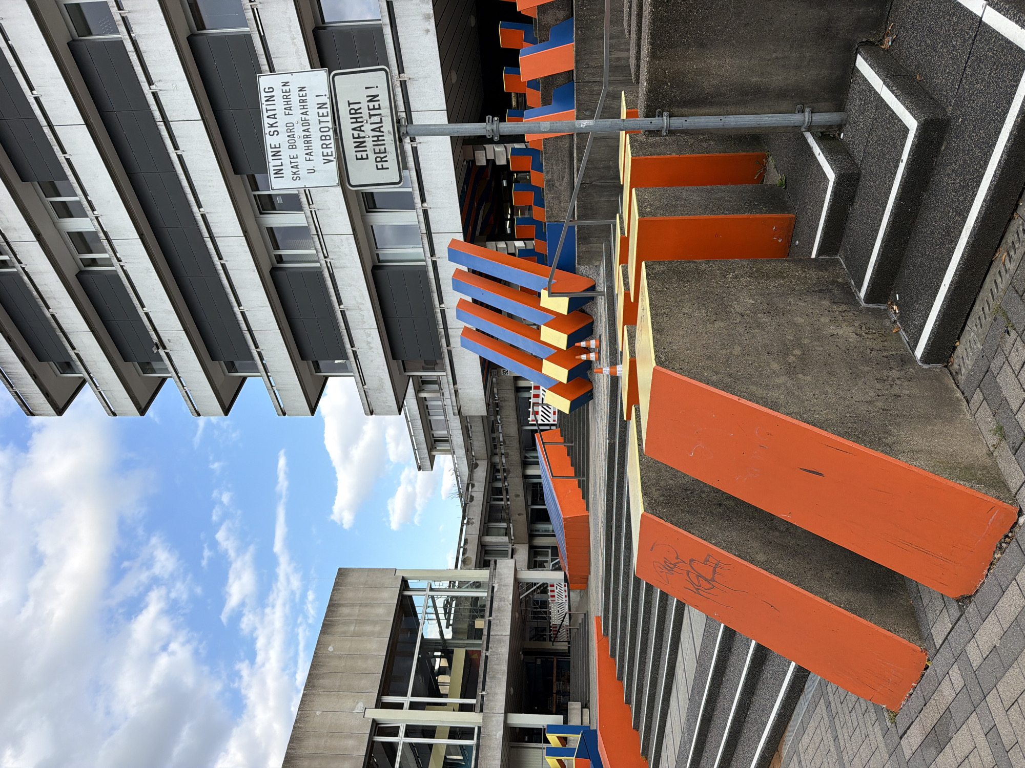 The image depicts an outdoor area of a modern building complex, likely a public space or educational institution, located in Schwelm, Germany. The foreground features a concrete staircase with a textured, dark gray surface and white contrasting edges. Adjacent to the stairs are angled concrete structures painted in bright orange, with some exhibiting graffiti. Further into the scene, there are more concrete structures and a larger staircase leading up to a multi-story building. The building itself is constructed of concrete and glass, with dark gray horizontal paneling on its upper floors. Two white signs are prominently displayed on a metal pole in front of the building. The top sign reads "INLINE SKATING SKATE BOARD FAHREN U. FAHRRADFAHREN VERBOTEN!" (Inline skating, skateboarding, and cycling forbidden!). The bottom sign states "Einfahrt FREIHALTEN!" (Keep entrance clear!). In the background, there are abstract, colorful geometric sculptures in orange and blue. The sky above is blue with scattered white clouds, and the overall environment suggests a weekday with no people visible.