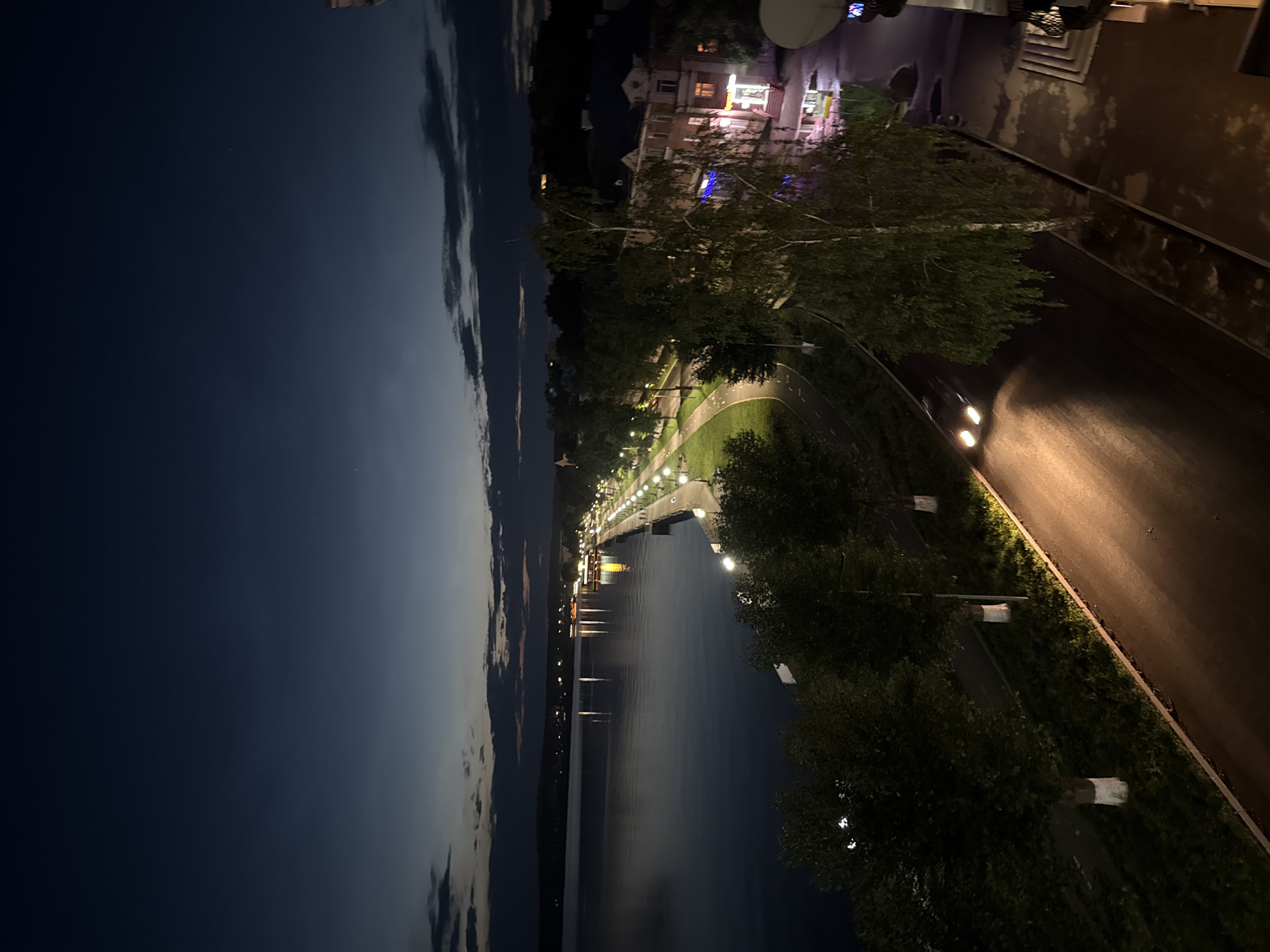 The image shows a waterfront promenade and adjacent street in Votkinsk, Russia, at twilight or early night. The water body, likely the Votkinsk Reservoir, is calm and reflects the lights of a distant embankment lined with streetlights and buildings. A paved walkway runs along the waterfront, bordered by trees and grass. Beyond the walkway is a road with a single car driving, its headlights illuminating the asphalt. On the right side of the road, there are buildings with illuminated windows and signs. The sky is a deep blue with a few faint clouds. The overall scene depicts a peaceful urban evening by the water.