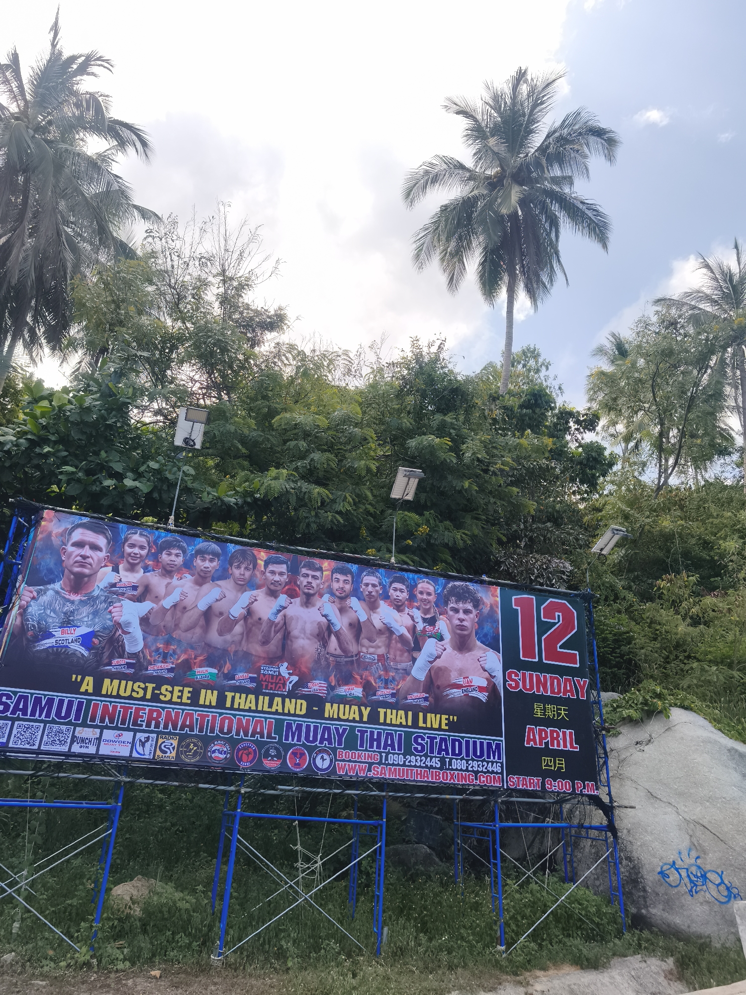 The image depicts a large advertisement billboard for the "Samui International Muay Thai Stadium", located in Baan Chaweng Noi, Thailand. The billboard features images of several Muay Thai fighters and advertises a "Muay Thai Live" event, branded as a "must-see in Thailand". It lists the date as "Sunday, April 12" with the start time at 9:00 P.M. The billboard is mounted on scaffolding, with lush green vegetation and palm trees visible in the background. A large boulder with graffiti is to the right of the billboard. Above the billboard are three small solar panel lights.