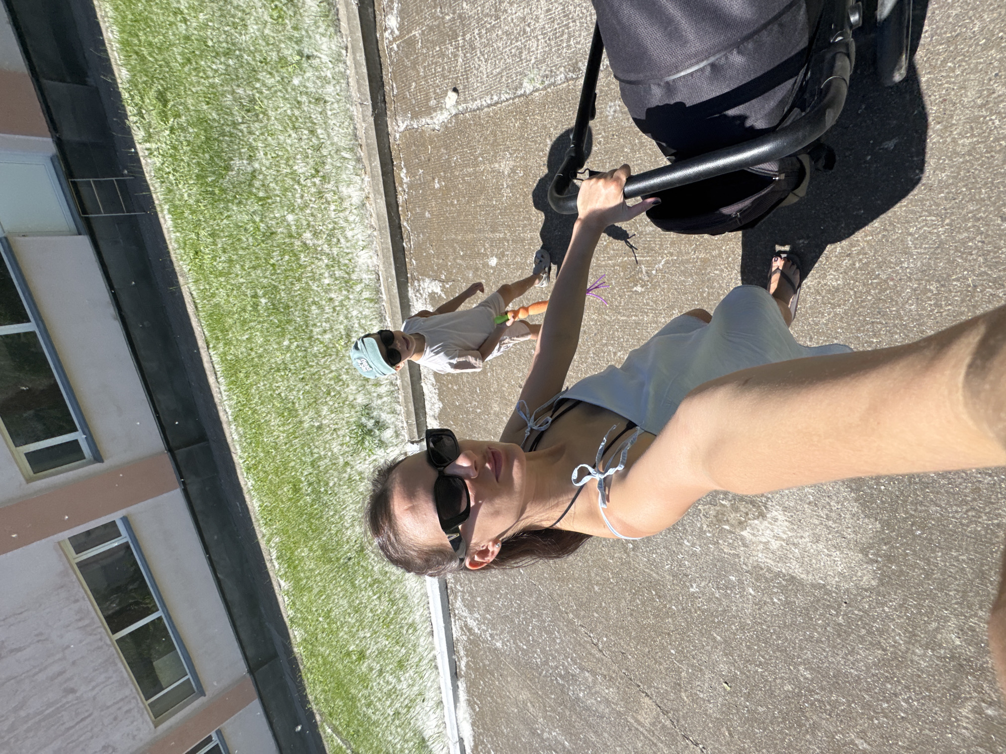 In Chaikovsky, Russia, a woman wearing a light blue dress and black sunglasses is taking a selfie outdoors. She is pushing a black stroller and walking on a concrete path. To her left, a patch of green grass is visible, covered in white fluff. A young boy, wearing a light-colored t-shirt, shorts, sunglasses, and a blue baseball cap, stands on the grass holding an orange toy and a purple string. He is facing away from the camera. In the background, a light-colored building with multiple windows is visible. The scene suggests a warm, sunny day, possibly during a season with poplar trees shedding their fluff.