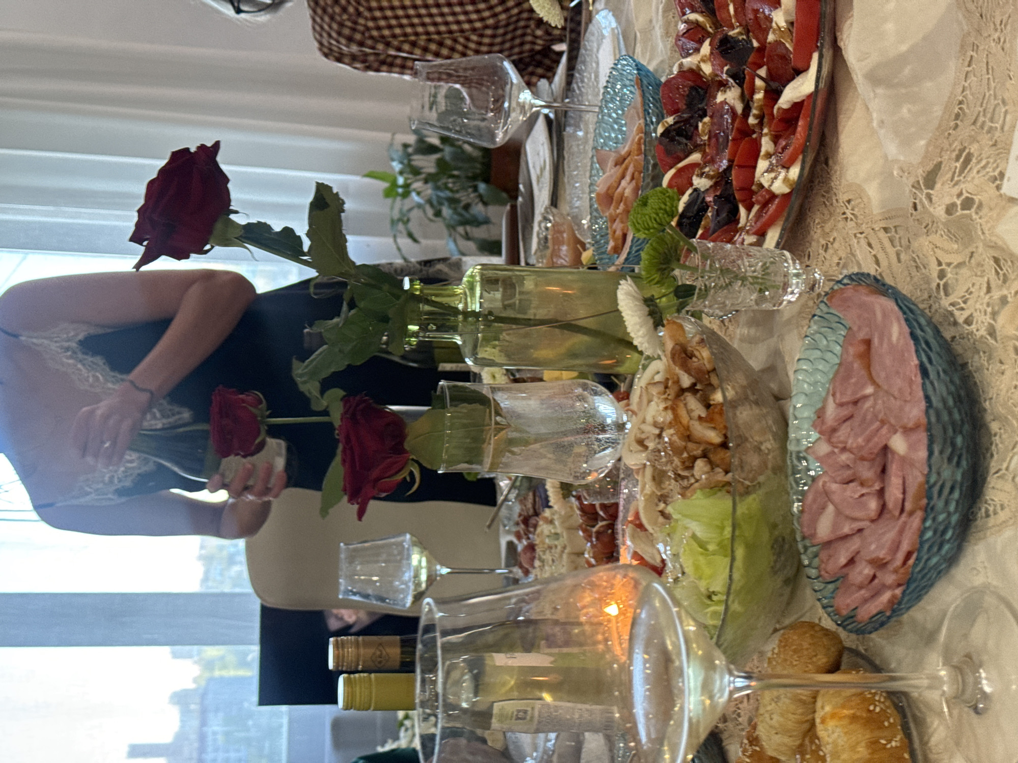 A photograph shows a table set for a celebratory meal. The table is covered with a light-colored lace tablecloth. On the table are several glass vases containing red roses, various plates of food including what appears to be caprese salad and sliced meats, and several wine glasses. Two wine bottles are partially visible near the bottom of the frame. A person wearing a black top with lace detailing is seen holding roses. The background shows an out-of-focus urban landscape, suggesting a city location. The scene is indoors, likely a residential space given the furniture and decor. The location is provided as Krasnoyarsk, Russia.