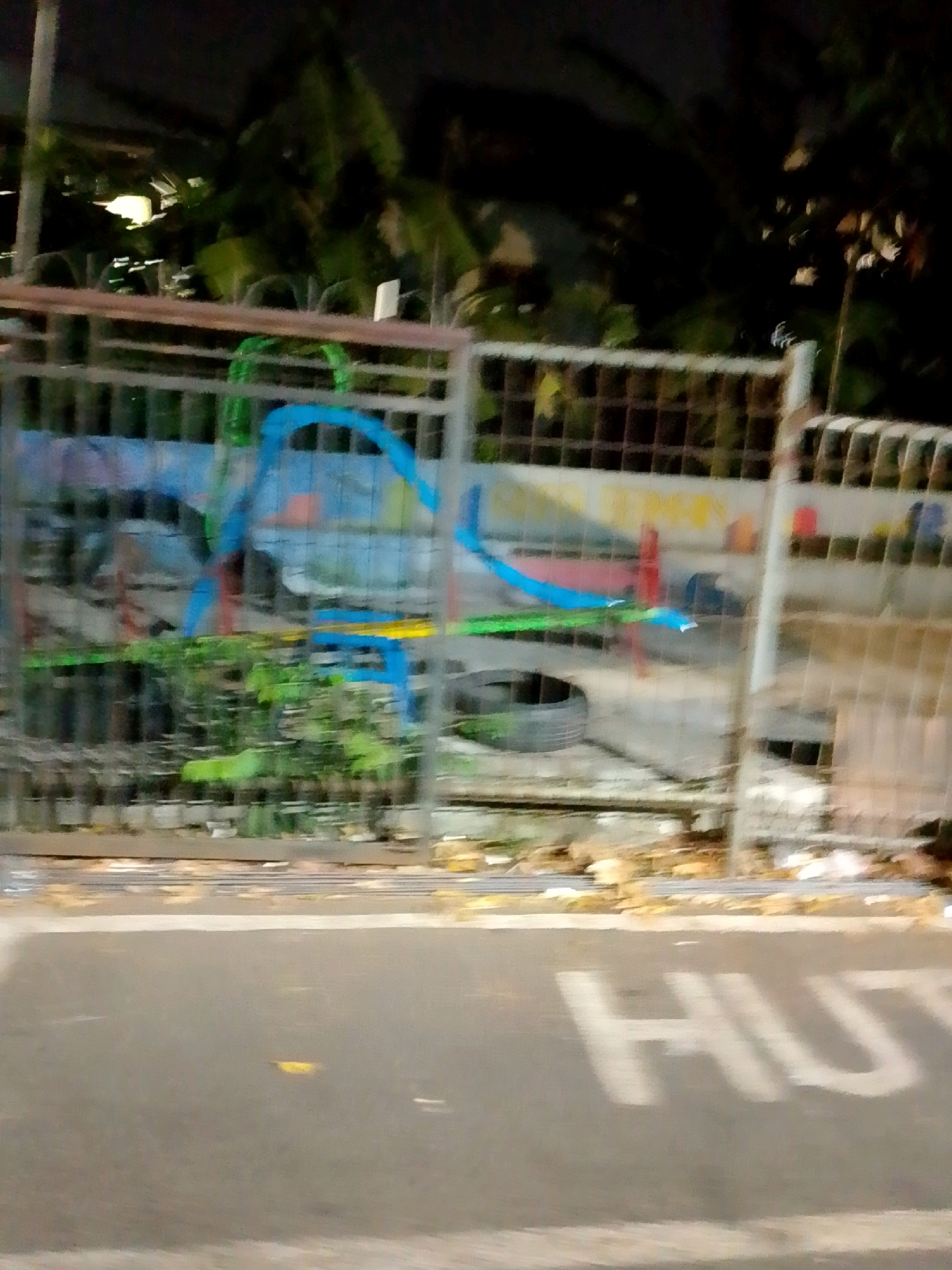 The image is a blurry, low-light photograph of a street scene in East Jakarta, Indonesia. In the foreground, a paved road shows a faint white marking that appears to spell "HUT" or similar letters. Behind the road, a metal fence runs horizontally, obscuring a cluttered area beyond. This area contains graffiti on a concrete wall, with prominent blue and yellow spray paint. A green plastic structure, possibly part of a playground, is visible behind the fence. Several tires and debris are scattered in the background, suggesting a neglected or construction site. Lush green foliage is visible above the fence, indicating the presence of trees and other plants. The overall impression is of an urban environment at night, with limited visibility and a sense of disarray.