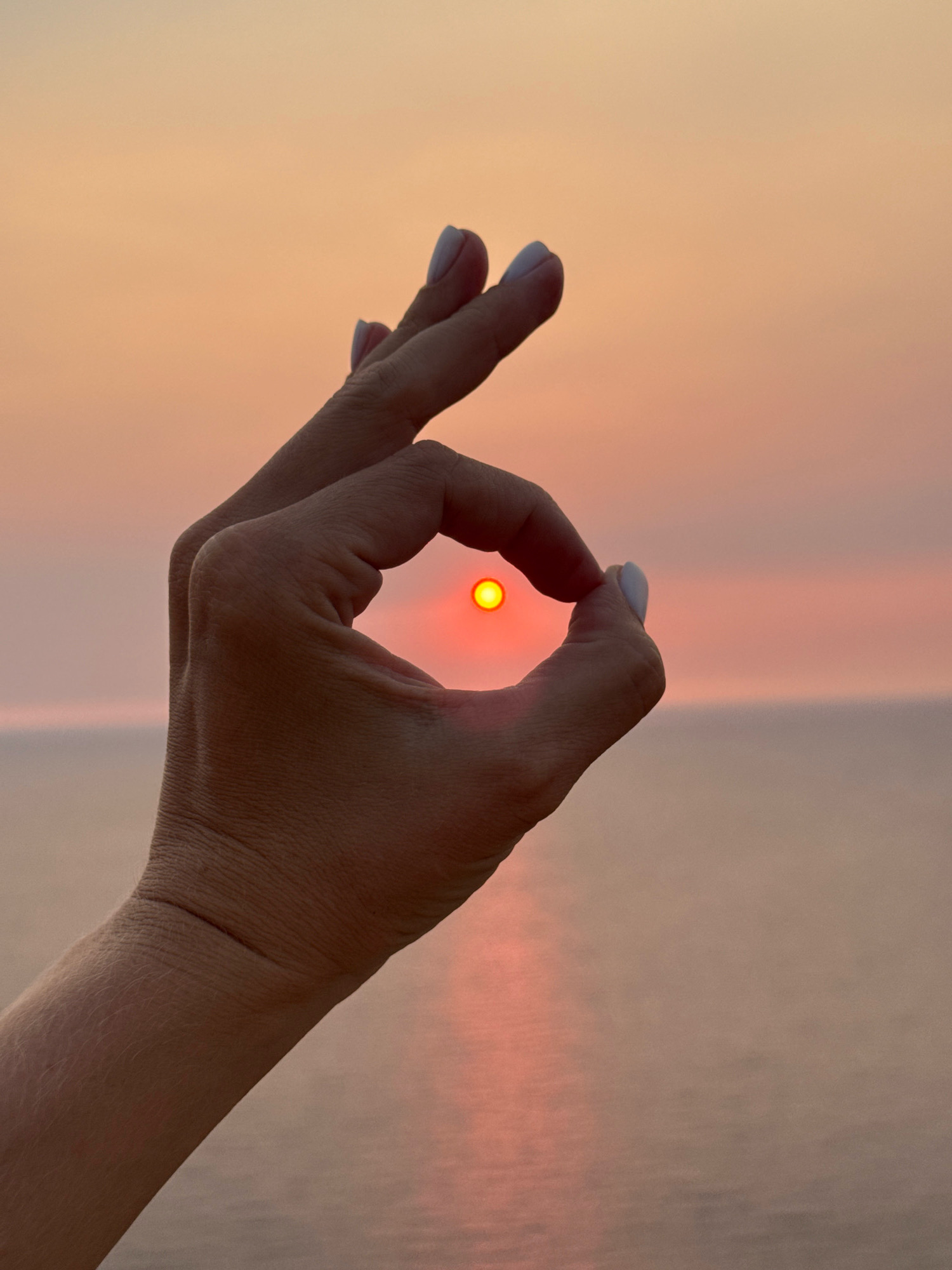 An extreme close-up shot focuses on a person's right hand, with the fingers forming an "okay" symbol. The sun, a small, bright orange orb, is perfectly framed within the circle created by the thumb and index finger. The backdrop is a soft gradient of sunset colors, transitioning from pale orange at the top to a muted pink and then a hazy grey-pink near the horizon. The water, likely the sea, is visible in the lower half of the image, reflecting the warm hues of the sky. The image does not provide any information about the city of Zagora, Montenegro, or any specific location.