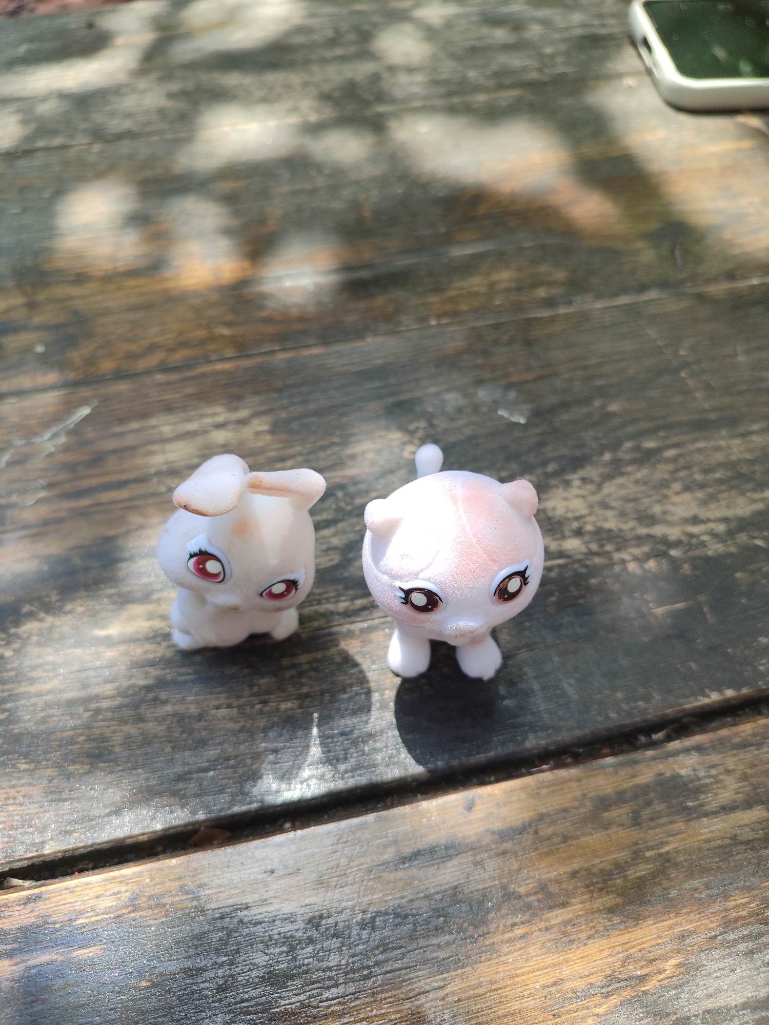 Two small, light-pink toy figurines, one resembling a rabbit and the other a cat, are positioned on a weathered, dark wooden surface. The rabbit figurine, on the left, has long ears and large, red-rimmed eyes with black pupils. The cat figurine, on the right, has short ears, a tail, and large, brown-rimmed eyes with black pupils. The wooden surface appears to be made of planks with visible grain and wear. Sunlight casts shadows and dappled light across the scene. A corner of a white smartphone is visible in the upper right of the frame. The setting appears to be outdoors, possibly a patio or deck, in an unidentified urban location.