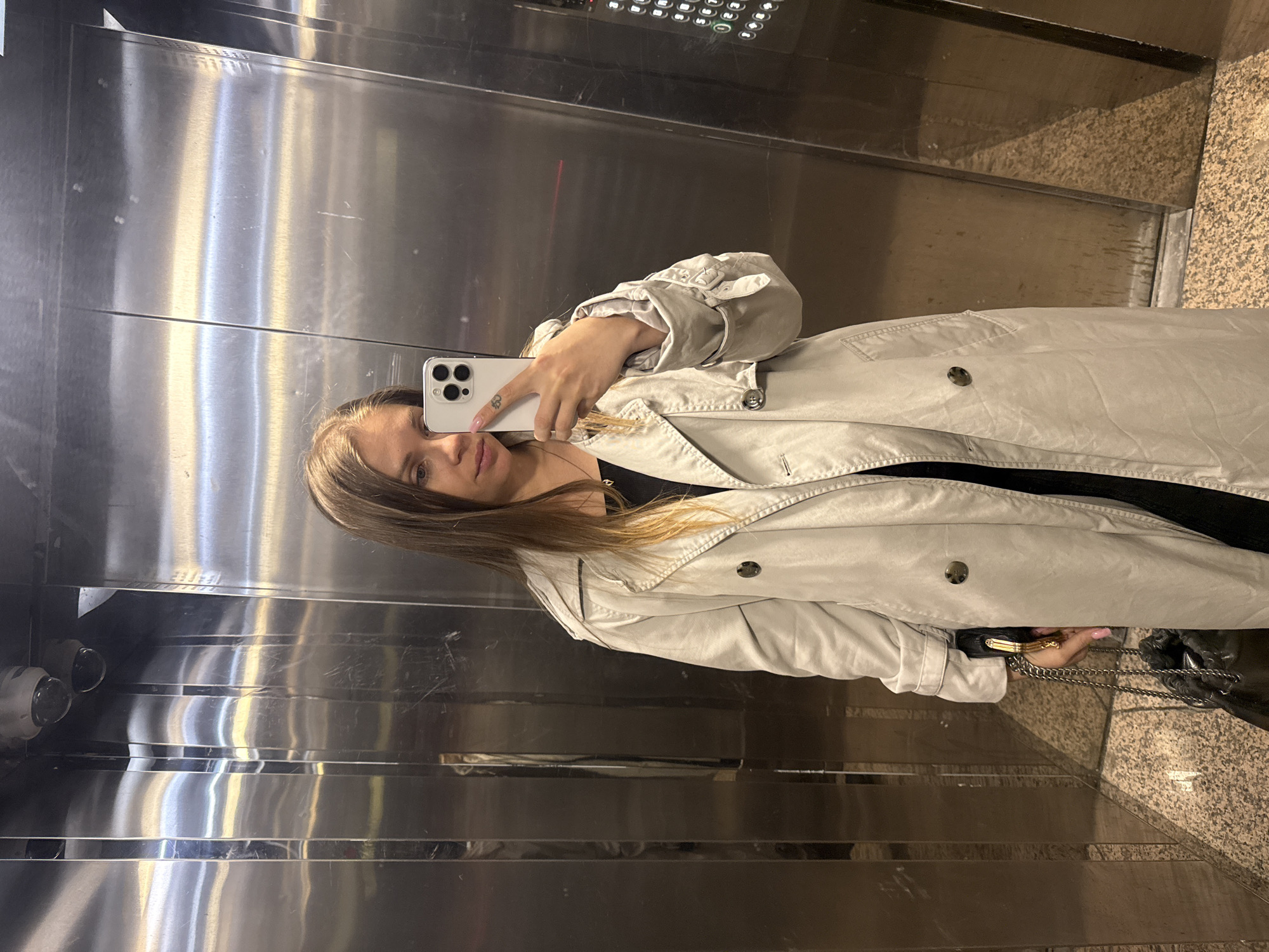 A young woman is depicted in a mirror selfie taken inside an elevator. She is positioned slightly to the left of the frame, facing the camera with a neutral expression. She has long, straight brown hair with lighter ends and wears a light beige or light grey trench coat over a dark top. Her right hand, with light pink nail polish and a small, dark tattoo on her index finger, holds a white smartphone with a triple-lens camera system, indicating she is taking the photograph. Her left hand, partially visible near her hip, holds a black handbag with a metallic chain strap and a gold-colored clasp.
The setting is an elevator interior, featuring highly reflective, brushed stainless steel walls that show reflections of the woman and various elements. On the upper right, a control panel with illuminated buttons is visible in the reflection, though the numbers on the buttons are not clearly legible. A faint red light streak can be seen below the control panel. On the left side, two circular lenses, likely part of a security camera, are visible in the reflection. The floor of the elevator is a light-colored, speckled stone or granite. The lighting is artificial, typical of an indoor environment. The scene takes place in Batumi, Georgia.