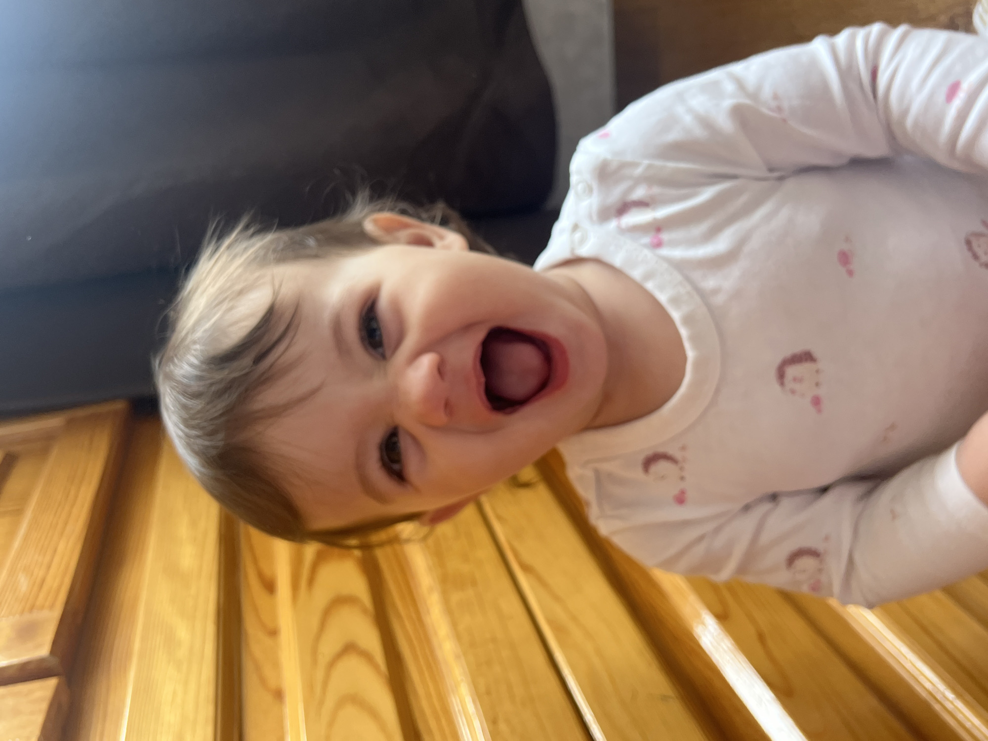 A baby with light brown hair is positioned off-center in the frame, with a wide, open mouth and visible teeth. The baby is wearing a white, long-sleeved shirt patterned with small, pink human-like figures. The baby's head is tilted to the left. The background includes a dark brown object that appears to be a couch and a light brown wood paneling. The location, Elena, Bulgaria, is not evident in the image.