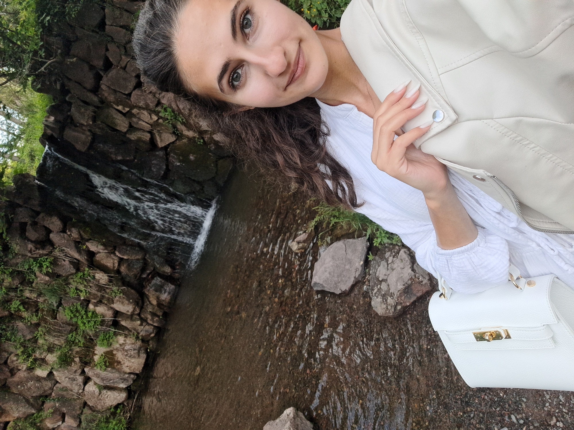 This is a vertical, slightly tilted selfie of a young woman in Lokorsko, Bulgaria. The woman has long, dark, curly hair, and is looking directly at the camera with a gentle smile. She is wearing a white, loose-fitting shirt and a light-colored jacket, possibly leather. A white handbag with gold hardware is visible in the lower right corner.
The background features a small waterfall cascading over a wall of dark rocks interspersed with patches of green vegetation. The water flows into a shallow, rocky stream. The overall setting appears to be natural and serene, possibly a park or a scenic outdoor area.
The lighting suggests it is daytime, and the natural environment indicates an outdoor setting. The woman's relaxed posture and smile convey a sense of contentment and enjoyment of the moment. There is no visible text in the image.