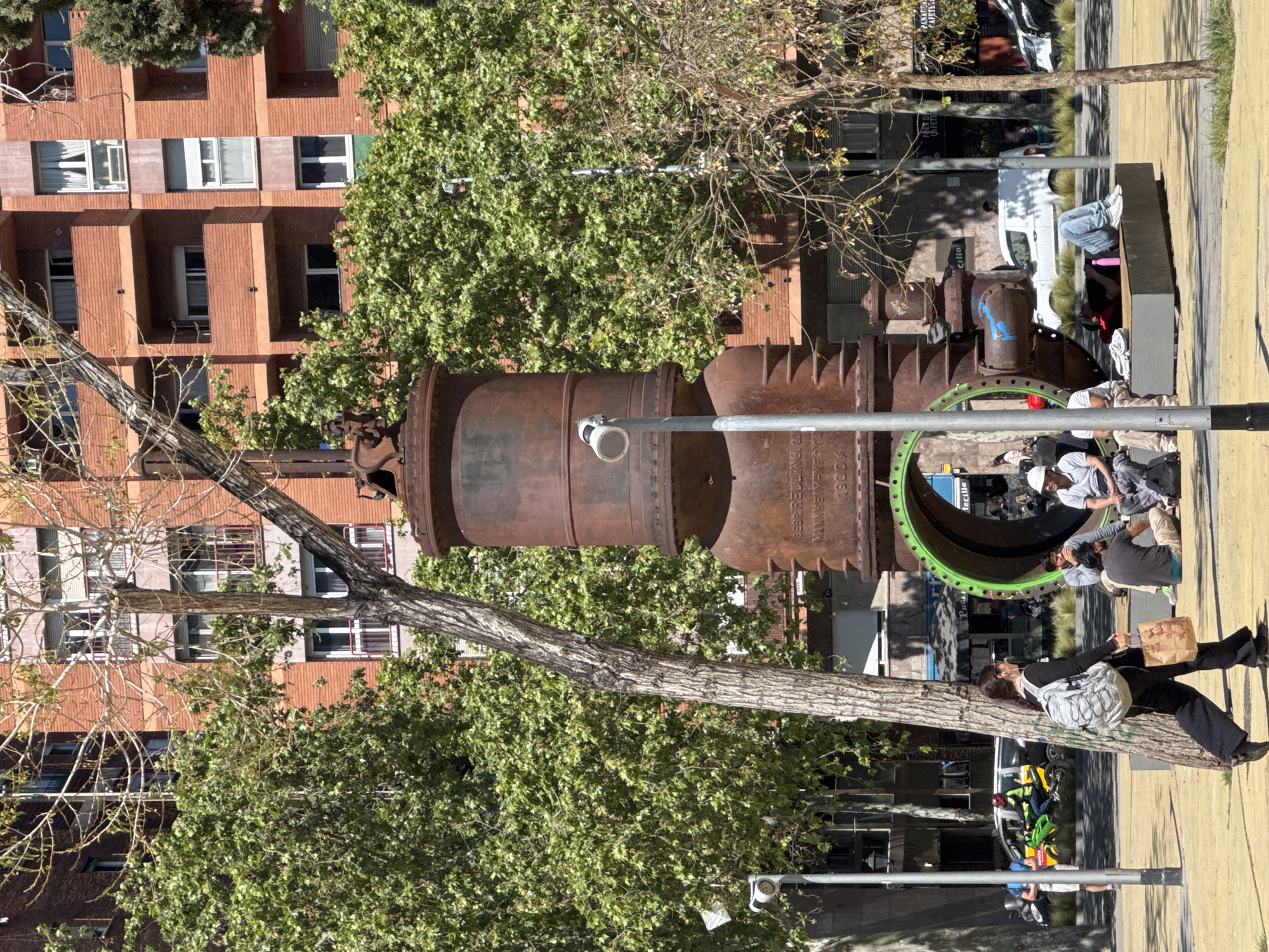 This image captures a vibrant urban scene in Barcelona, featuring a large, rusty industrial structure that appears to be repurposed public art, alongside people engaged in leisure activities. The central focus is a monumental, cylindrical metal object, heavily rusted, with intricate details. Visible text on its surface includes "GEBR. REL... M. & BANTIN" and "1900," suggesting its historical origin or manufacturing details. One end of this structure reveals a striking green-painted interior rim, contrasting with its aged exterior.
Several individuals are present in the bright daylight. To the right, a person in blue jeans and a dark top is seated on a concrete bench. Near the green-rimmed pipe, a small group of people are gathered, including one wearing a white cap, appearing to socialize or relax. In the immediate foreground, a woman with dark hair and a light-colored backpack leans casually against a tree trunk, holding a paper bag.
The background features a multi-story building constructed from reddish-brown bricks, characterized by numerous windows and recessed, square-shaped balconies. Green foliage from several trees adds natural elements to the urban setting, while some trees also show bare branches. The ground consists of distinct yellow paving alongside darker asphalt, with vehicles, including a white van, visible further in the distance. A lamppost stands in the foreground, and shadows indicate a sunny day, creating a sense of everyday life in a public park or plaza.
