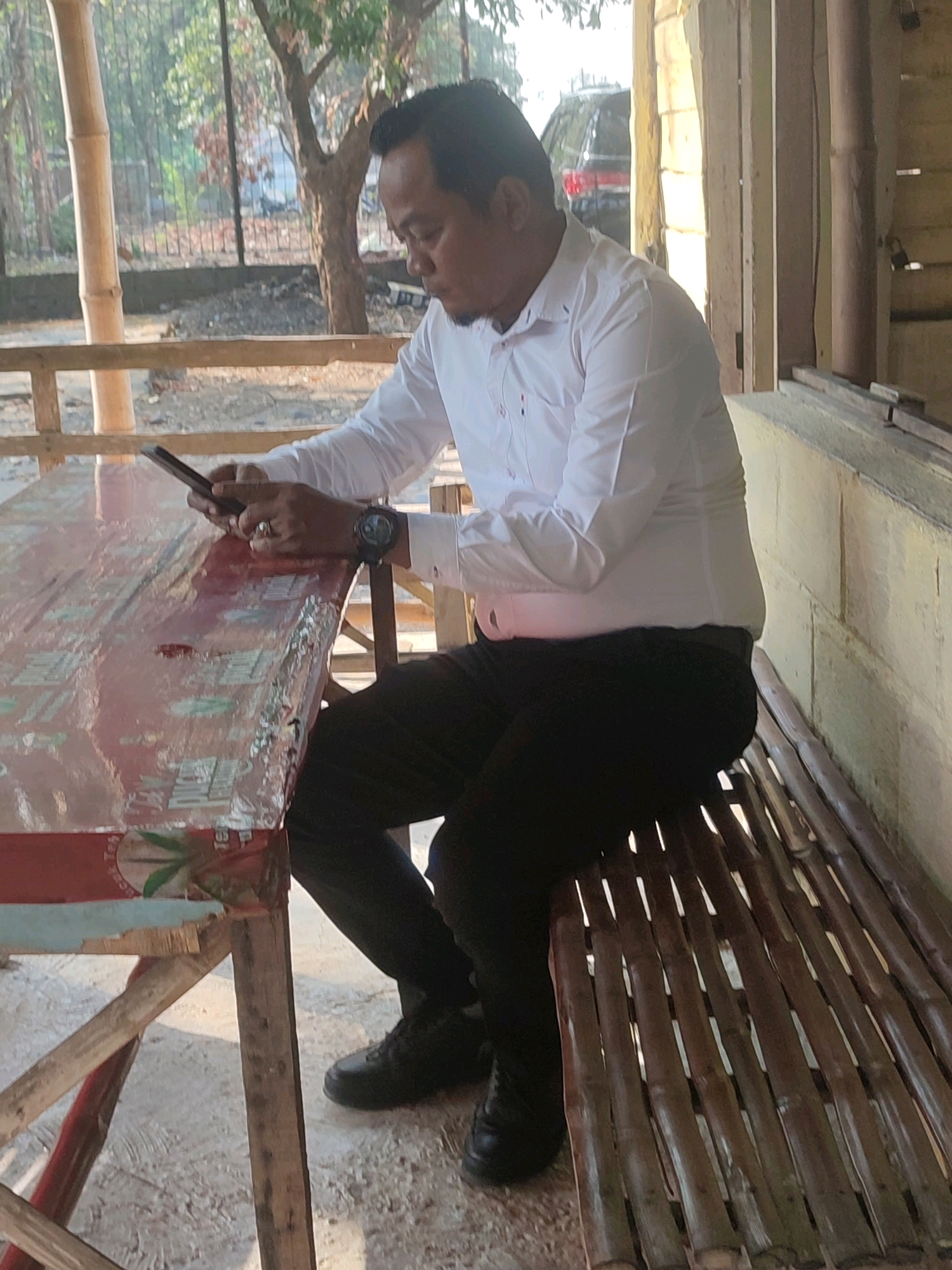 In Ringinanom, Indonesia, a man is seated at a table covered with a red cloth, holding a smartphone. He is wearing a white long-sleeved shirt, black pants, and black shoes. A watch is visible on his left wrist. The scene appears to be outdoors, with a bamboo fence and trees in the background, along with a dark-colored vehicle. The table has a rustic design, and the seating appears to be made of bamboo. The ground is a light-colored, granular surface.