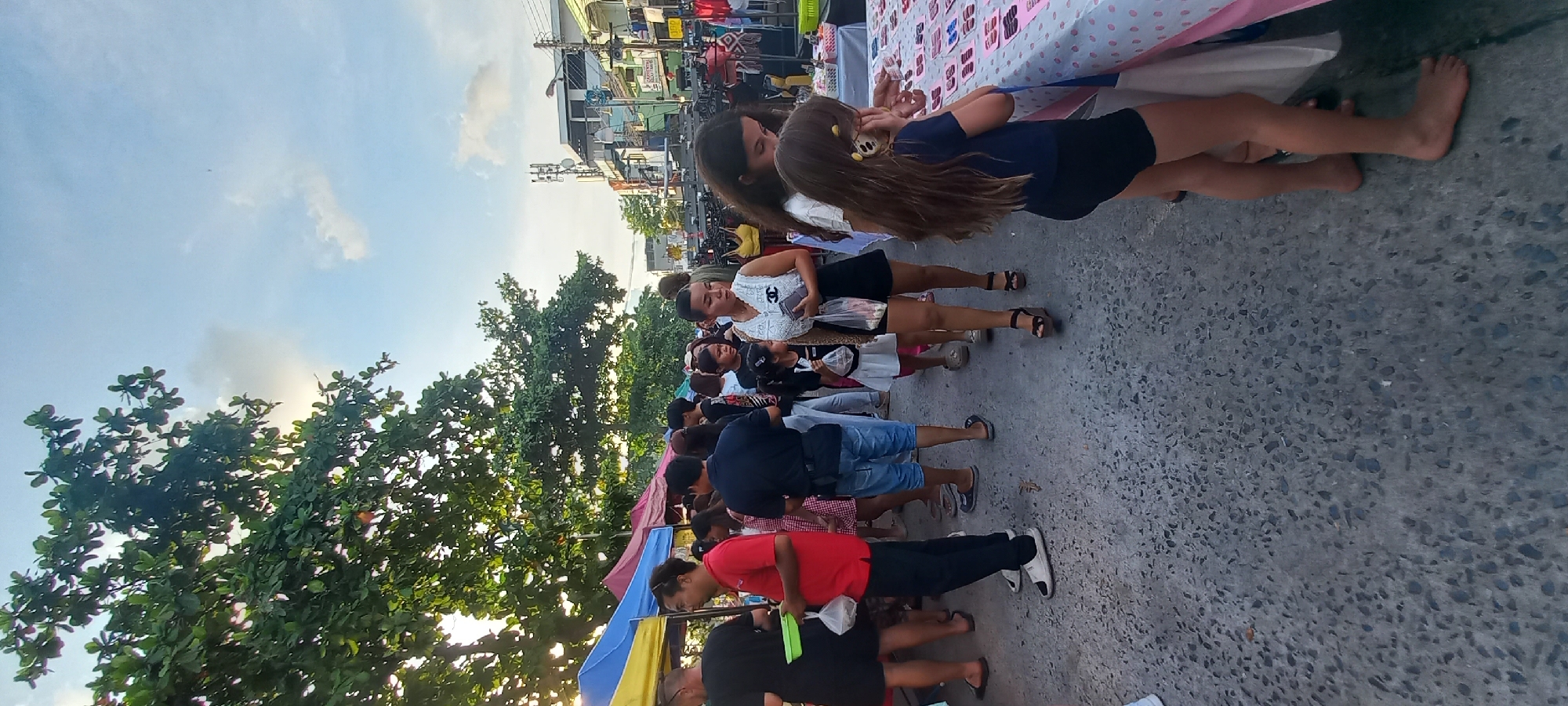 The image depicts a street market scene in Thong Sala, Thailand, crowded with people browsing various stalls. A line of individuals is visible, including a child with long hair wearing a blue shirt and black shorts, standing next to a woman in a white top and black skirt. Many individuals wear casual clothes. The foreground shows a paved street surface, while the background features trees, buildings, and stalls covered with fabrics. The sky is visible in the upper portion of the image.